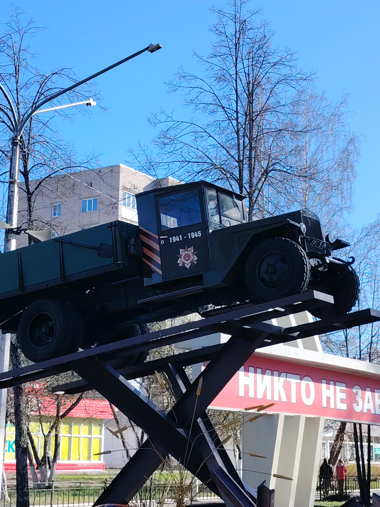 The image depicts a military truck mounted on a steel monument in what is described to be the city of Chaikovsky, Russia. The truck is dark green, and has a display on its side indicating the years 1941-1945, likely referencing World War II. The monument beneath it is made of black steel beams intersecting in a crisscross pattern. The entire structure rests on a concrete base with a red and white banner that bears a message in Russian. In the background, there is a multi-story building with light brick and windows, along with leafless trees against a bright blue sky. A street light is also visible on the left side of the frame. Parts of other buildings and street scenes are visible in the background, including pedestrians.
