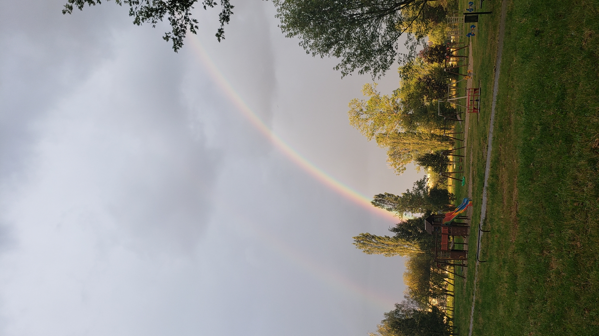 The image captures a park scene in Prešov, Slovakia, dominated by a cloudy sky and a prominent double rainbow. The foreground features a grassy area with a paved pathway winding through it. In the middle ground, a children's playground is visible, complete with swings, a slide, and other play structures. A variety of trees, including tall, slender ones and more mature, leafy ones, are scattered throughout the park. The background shows a line of buildings, suggesting the park is situated within an urban or suburban area. The lighting indicates it may be late afternoon or early evening, with the overcast sky casting a diffused light over the scene.
