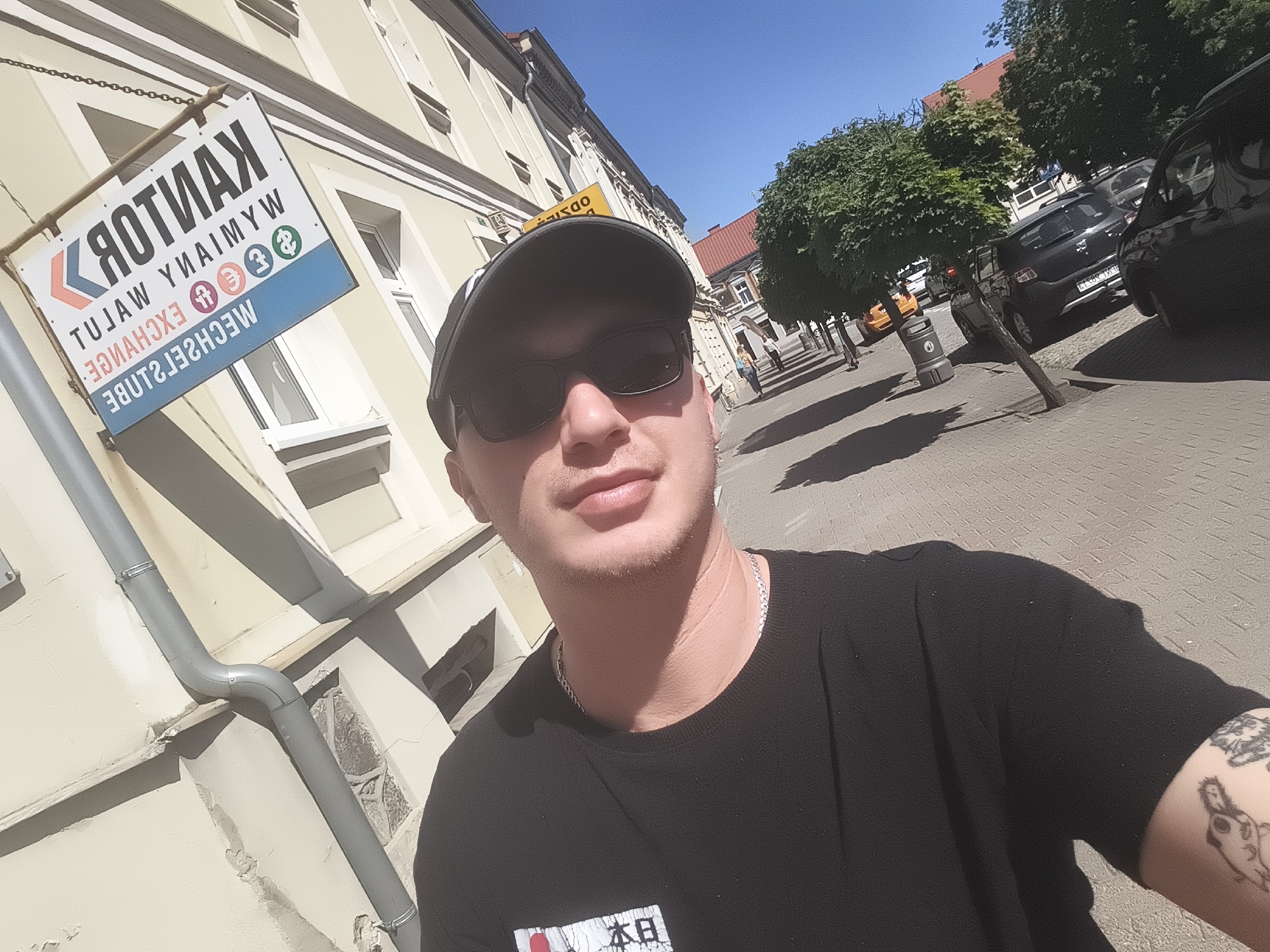 The image is a selfie taken by a man wearing sunglasses and a black baseball cap in Nowy Tomyśl, Poland. The man is wearing a black t-shirt and a silver necklace. He has tattoos on his right arm.
Behind him is a building with a "Kantor Wymiany Walut" (Currency Exchange) sign. The sign is in Polish and lists currencies such as the euro, dollar, and pound. A downspout is attached to the building.
The background shows a street with trees, parked cars, and pedestrians in the distance. The sky is blue and sunny. The street appears to be brick or stone paved.