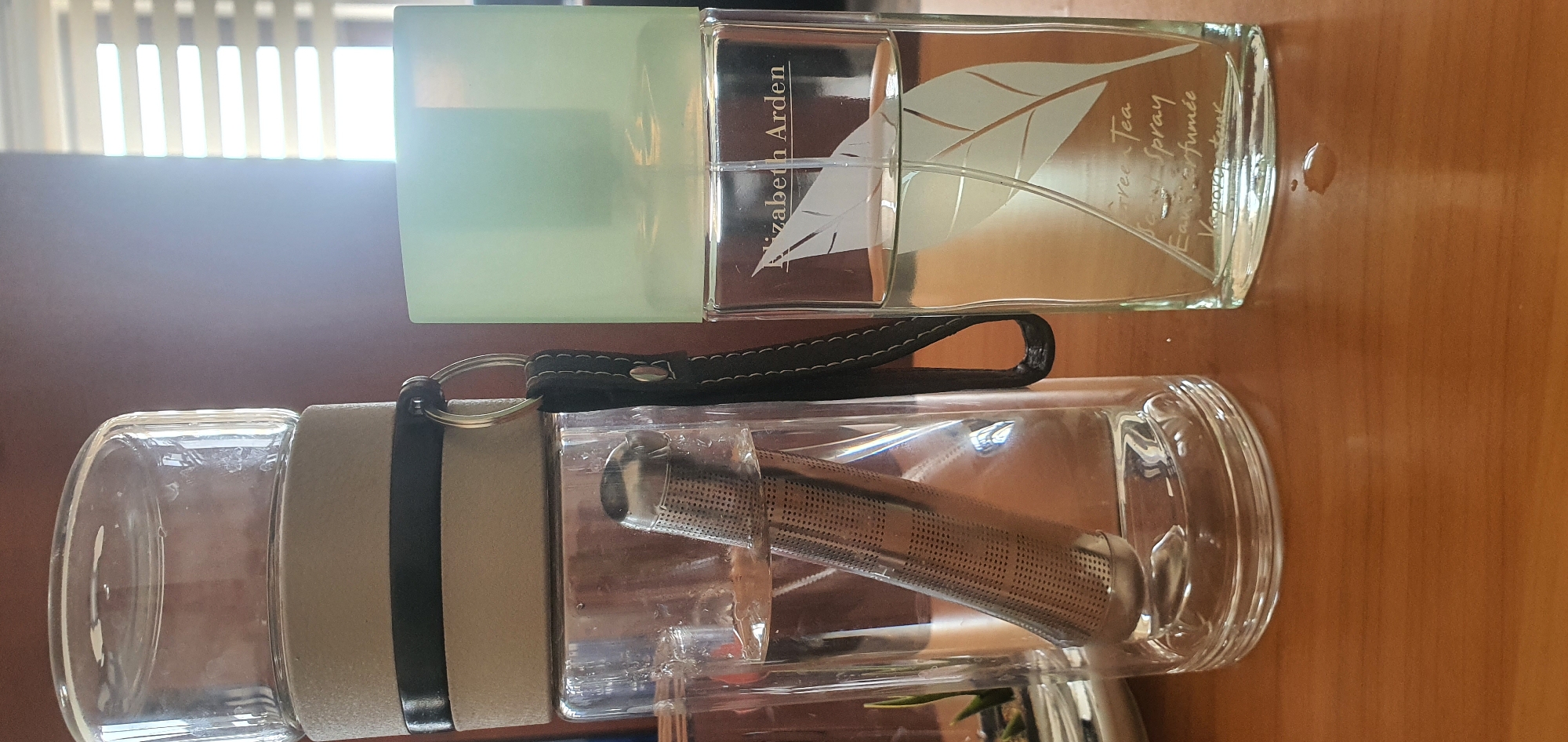 The image shows two glass containers on a wooden surface, possibly a desk. On the left is a clear glass cylinder with a silver tea infuser submerged in clear liquid. The cylinder is topped with a beige and black sleeve, and a black strap with a silver ring is attached to the sleeve. To the right is a clear glass perfume bottle with a pale green cap. The bottle contains a pale yellow liquid and has a delicate leaf design etched onto it. Text on the bottle reads "Elizabeth Arden Green Tea Scent Spray Eau parfumee Vaporisateur". The background is a blurred brown wall and a window with horizontal blinds. The lighting suggests it is daytime. The location is not specified in the image.