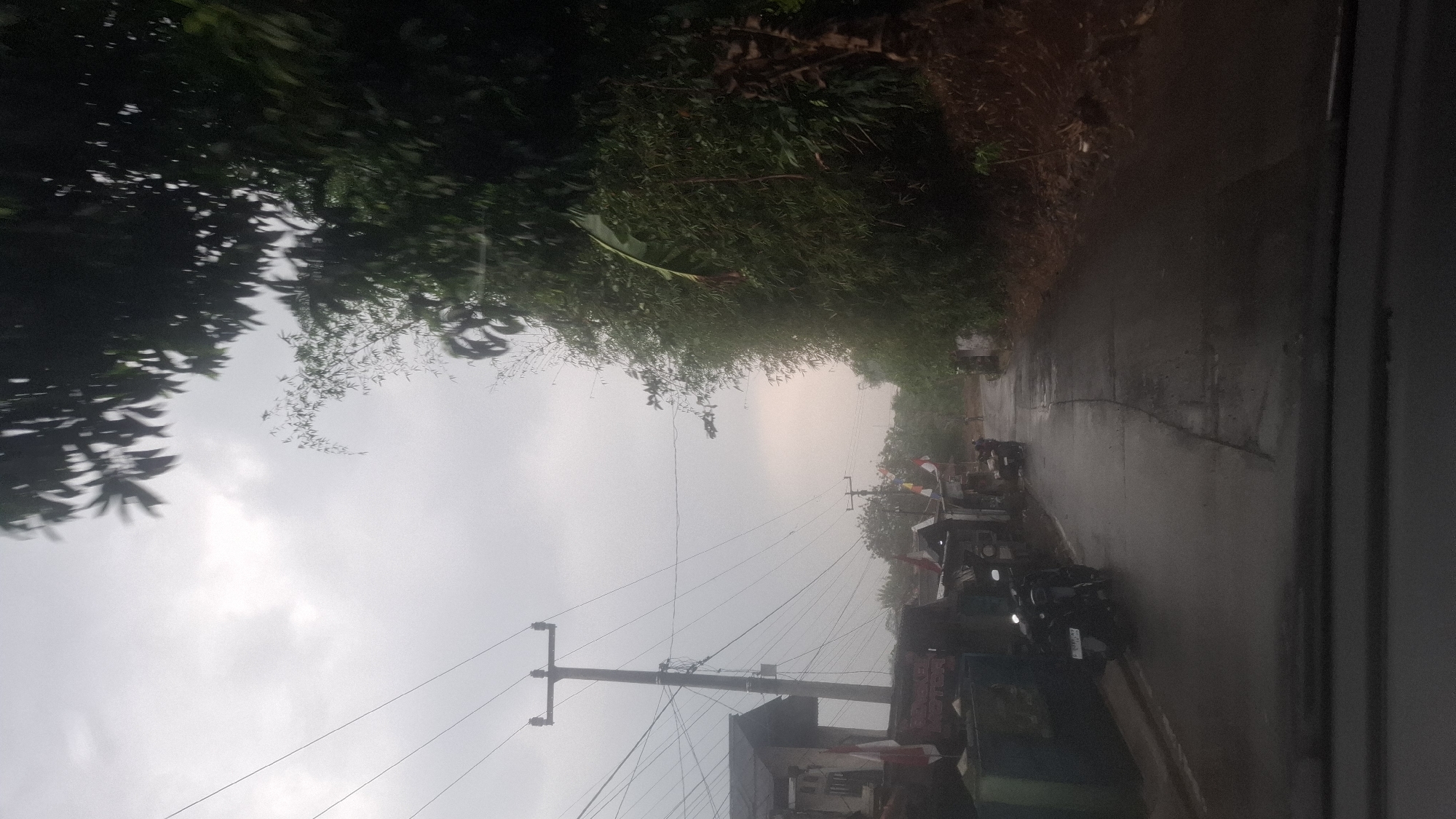 The image depicts a street scene in Sirnagalih, Indonesia, under a cloudy sky. A wet, asphalt road curves into the distance, bordered on the left by a few small buildings and on the right by dense, green foliage, likely bamboo and other trees. Utility poles with numerous wires are prominent on the left side of the road. Several motorcycles are parked along the roadside, and at least one person is visible walking further down the street. Indonesian flags are displayed on some of the buildings, suggesting a residential or commercial area. The overall atmosphere is overcast and damp, with the road appearing recently rained upon. The perspective suggests the photo was taken from inside a vehicle.