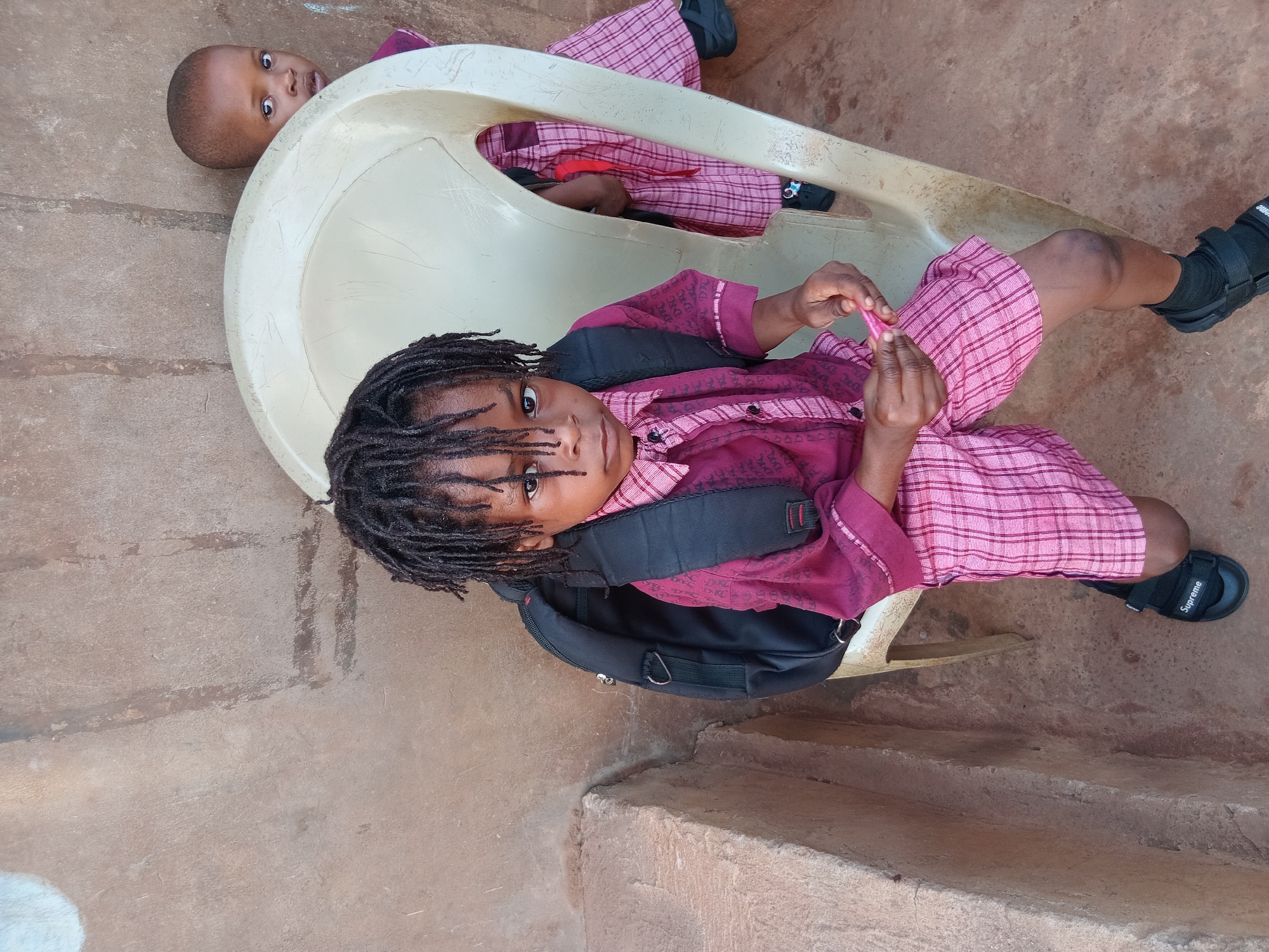 Two children wearing school uniforms are sitting on and near a light yellow plastic chair. The child in the foreground has dreadlocks and a black backpack. The child is wearing black sandals. The second child is positioned behind the chair. The setting appears to be an outdoor, perhaps on a porch or patio with a concrete floor and low wall. The location is Shagamu, Nigeria.