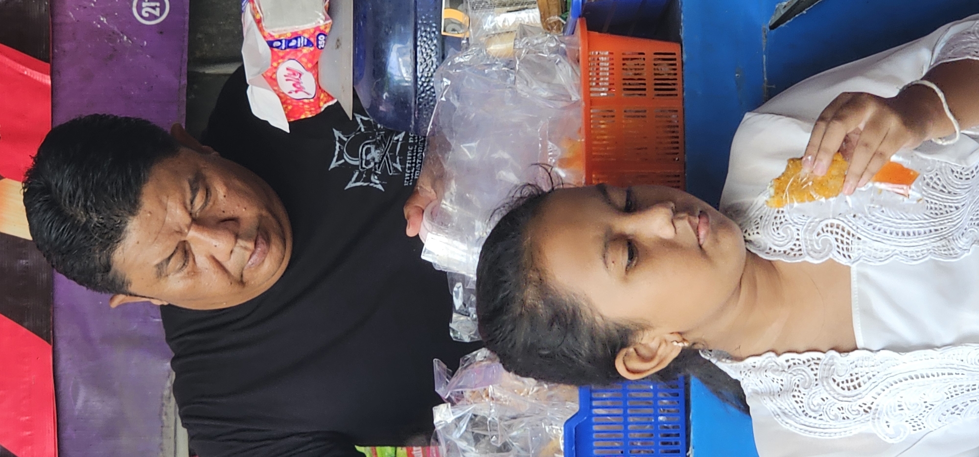 In Denpasar, Indonesia, a woman in a white lace-trimmed shirt is eating orange food. Next to her is a man wearing a black shirt with a white skull graphic. There is a container of tissues, multiple plastic bags, an orange basket, and a blue basket nearby.