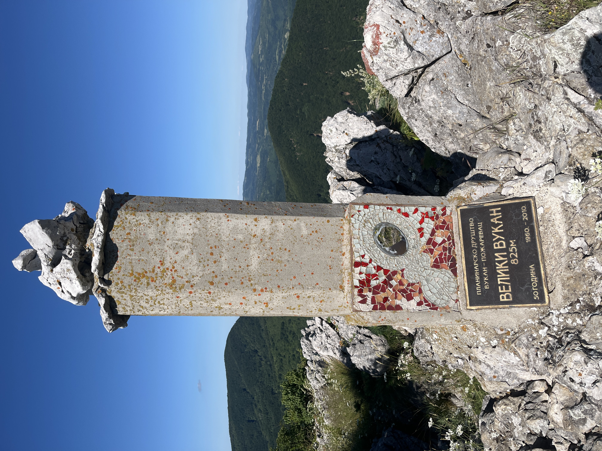 The image depicts the summit of Veliki Vukan, a mountain located in Petrovac-na-Mlavi Municipality, Serbia. A stone monument, topped with a stack of rocks, stands prominently in the foreground. The monument is adorned with a mosaic of red and white tiles and a circular reflective surface. Below the mosaic, a metal plaque displays text in Cyrillic script, identifying the location as "Veliki Vukan" and stating its elevation as "825M". The plaque also includes dates, "50 ГОДИНА" and "1960 - 2010". The monument is situated amidst a rocky, mountainous terrain with sparse vegetation. In the background, rolling, tree-covered hills stretch towards a clear blue sky. The overall scene is one of a scenic natural landscape, marking a significant point of elevation. There are no people visible in the image.
