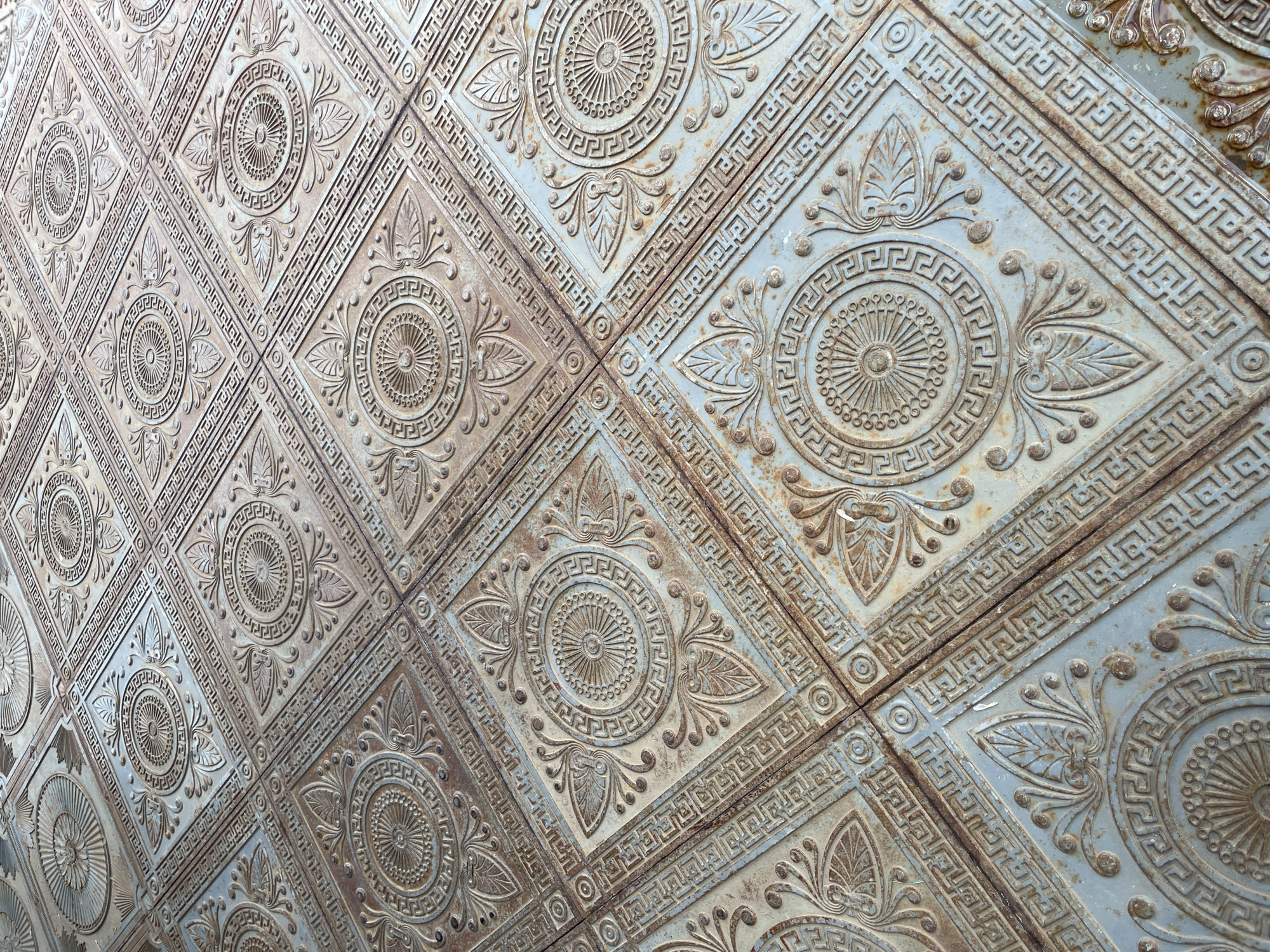 A close-up, high-angle shot captures an intricately patterned metal surface, likely a floor or decorative wall panel, located in Izhevsk, Russia. The surface is composed of numerous square or diamond-shaped tiles, arranged in a grid and viewed obliquely, creating a repeating geometric and floral design.
Each tile features a central circular motif with radiating lines, surrounded by a prominent Greek key (meander) pattern. Flanking this central circle are stylized, symmetrical floral or leaf designs. The borders of the tiles also incorporate Greek key patterns, creating continuous decorative lines across the entire surface.
The material appears to be aged metal, possibly cast iron or steel, characterized by a textured, slightly rough surface. The dominant colors are muted blues and greys, heavily intermixed with reddish-brown rust and darker patination in the recessed areas, indicating significant wear and exposure to elements. The overall impression is one of weathered elegance and historical design.
No people are present, and no specific activity or event is taking place; the image is a static portrayal of the decorative surface. The lighting is diffused, casting soft shadows that highlight the raised relief of the patterns. There is no discernible text. The weathered appearance suggests the surface is old or has been outdoors for some time.