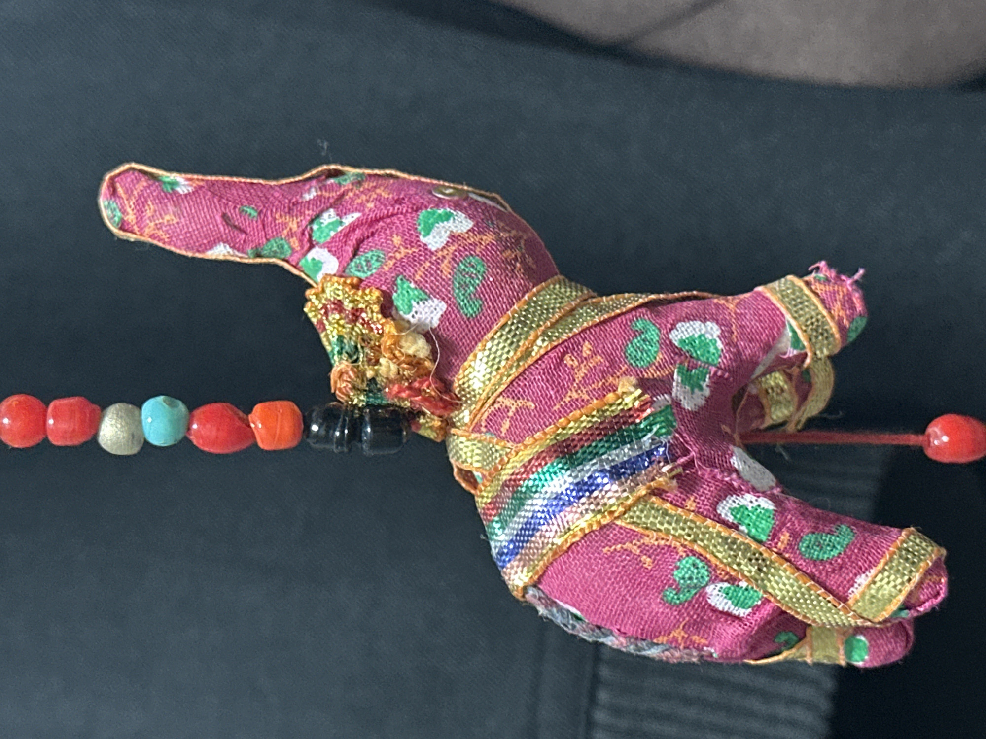 The image shows a close-up of a decorative fabric elephant ornament, likely a keychain or a hanging charm. The elephant is primarily pink with a repeating pattern of green and white floral or leafy designs. It is adorned with decorative gold trim and a colorful woven band around its body. Hanging above the elephant is a string of beads in various colors, including red, silver, turquoise, and black. A single red bead hangs from a red string below the elephant. The background is a dark, out-of-focus fabric, possibly clothing. There is no discernible location in the city of Nevinnomyssk, Russia.