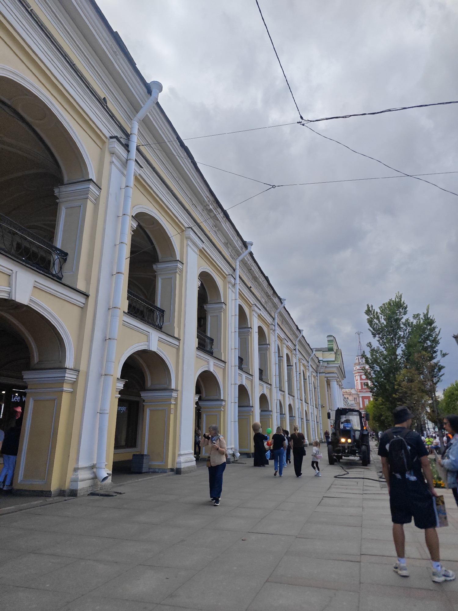 The image depicts a street scene in Saint Petersburg, Russia, likely on a cloudy day. The primary subject is a long, yellow building with a series of arched loggias and classical architectural detailing, including white pilasters and cornices. A white downspout runs vertically along the building's facade. Overhead, power lines crisscross the sky.
On the sidewalk in front of the building, several people are visible. Towards the left, two individuals are partially obscured by an entrance to a store. Further down the sidewalk, a woman in a light brown shirt and blue skirt holds a camera to her face. A small child in a pink dress walks near a black tractor, which has its headlights on and is connected to a hose. In the foreground on the right, a man wearing a black t-shirt, shorts, and a bucket hat faces away from the camera, carrying a bag. Another person in a denim jacket is to his right. In the background, a distinctive red and white clock tower with a spire is visible above the trees. The pavement is made of concrete slabs.