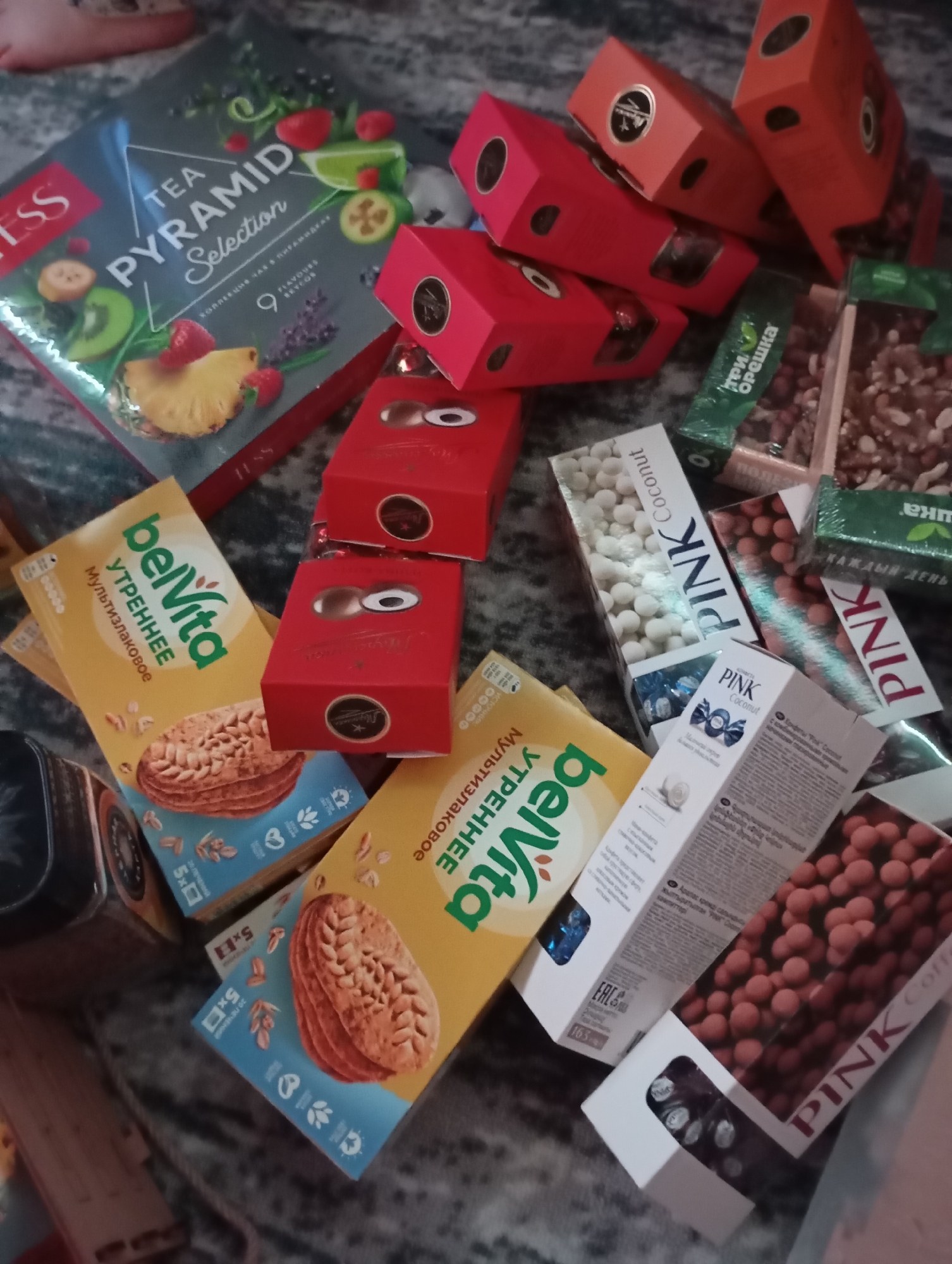 The image shows a collection of packaged food items scattered on a surface with a mottled gray and white pattern. A box of "TESS TEA PYRAMID Selection" is visible, along with several red boxes of confectionery, and boxes of "belVita" breakfast biscuits. Additionally, there are boxes of "PINK" candies, including "PINK Coconut" and "PINK Coffee" varieties, and a package of "3 nuts".