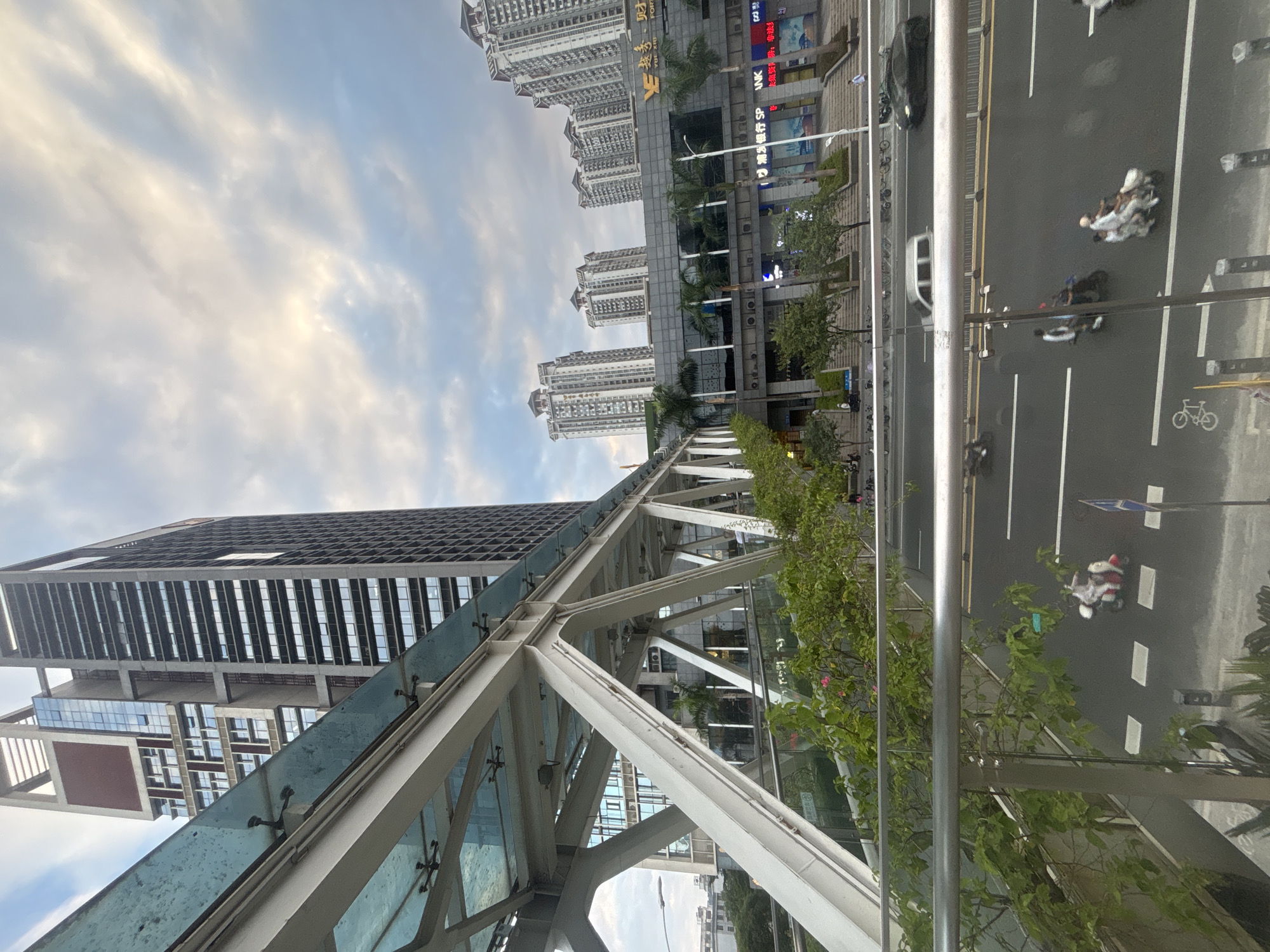 The image captures a street-level view of a city, likely in Liwan District, China, focusing on a modern pedestrian overpass with a glass and white metal structure. Below the overpass, a multi-lane road is visible with several vehicles, including scooters and a car, in motion. Greenery, including trees and bushes, lines the pedestrian walkway and the edge of the road. In the background, a tall, contemporary skyscraper with a dark facade and a prominent red square feature dominates the skyline, while several other high-rise apartment buildings with similar architectural styles are situated further back. The sky is partly cloudy with hints of sunlight breaking through. People are visible on the street, some riding scooters, and others walking. The overall scene depicts an urban environment with a blend of modern infrastructure and daily city life.