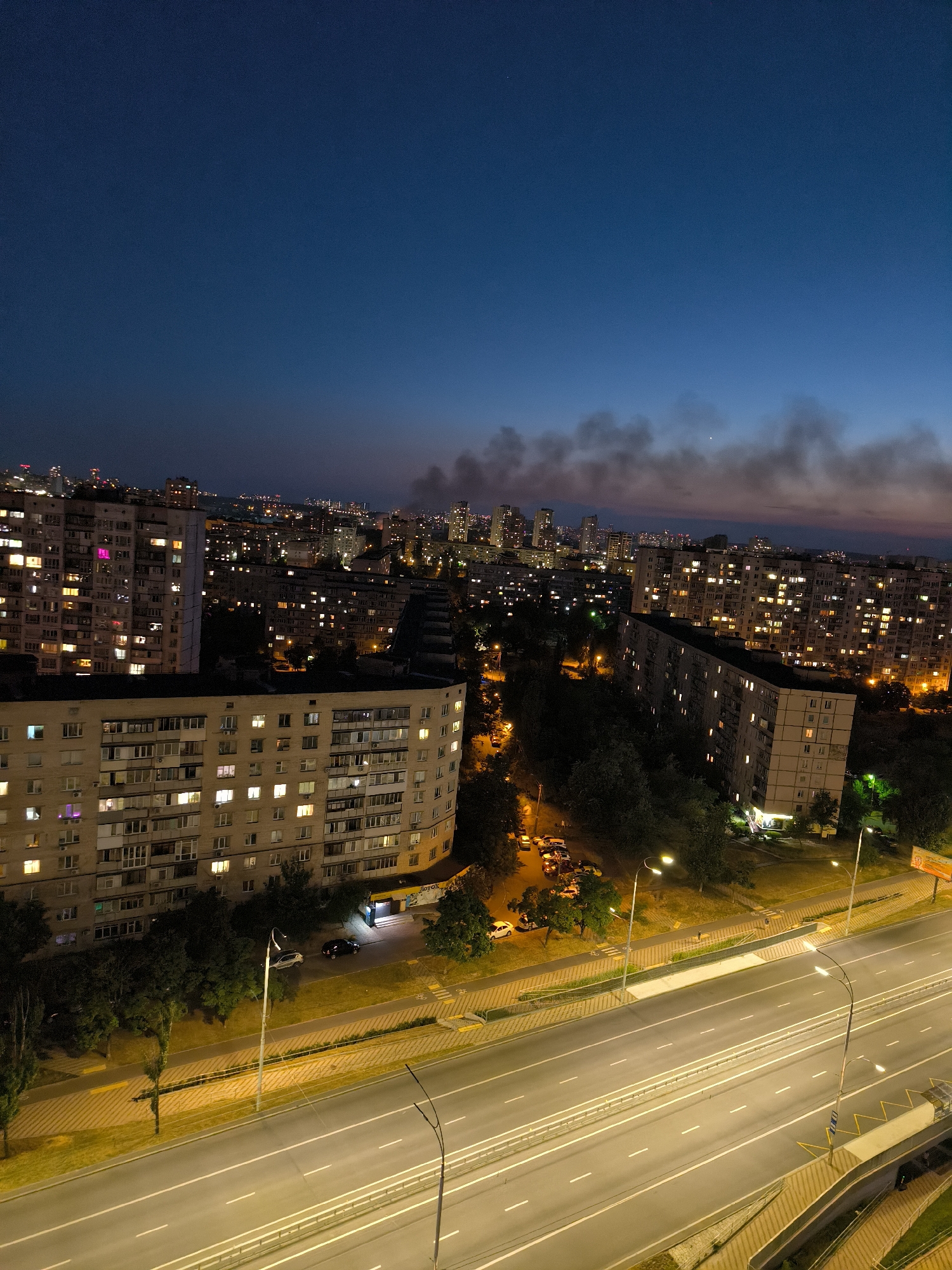 The image captures a nighttime aerial view of a residential area in Kyiv, Ukraine, featuring apartment buildings and a multi-lane highway. The sky is a deep blue, transitioning to lighter hues near the horizon, with wisps of smoke visible in the distance, suggesting potential atmospheric activity.
Dominating the foreground are several large, multi-story apartment buildings, their windows illuminated with a warm yellow light, indicating occupancy. Some buildings exhibit architectural styles common in post-Soviet urban planning, with multiple balconies and uniform facades. Trees are interspersed between the buildings and along the roadside, their dark foliage contrasting with the artificial lights.
A wide highway cuts across the lower part of the image. The road is illuminated by streetlights and the headlights of passing vehicles, which appear as streaks of light due to a long exposure, indicating motion. The highway has multiple lanes and is bordered by concrete barriers and sidewalks. Traffic appears to be moderate.
In the midground, a parking lot is visible next to one of the apartment buildings, with several cars parked. A streetlamp illuminates the area, casting a yellow glow on the vehicles and the surrounding pavement.
The distant cityscape is characterized by a dense collection of buildings, some of which are taller skyscrapers, their lights creating a sprawling urban panorama against the twilight sky. The overall impression is one of a bustling, active city at night, with the juxtaposition of residential areas, transportation infrastructure, and the natural elements of trees and sky. The presence of smoke in the distance adds a subtle element of intrigue to the otherwise typical urban night scene.