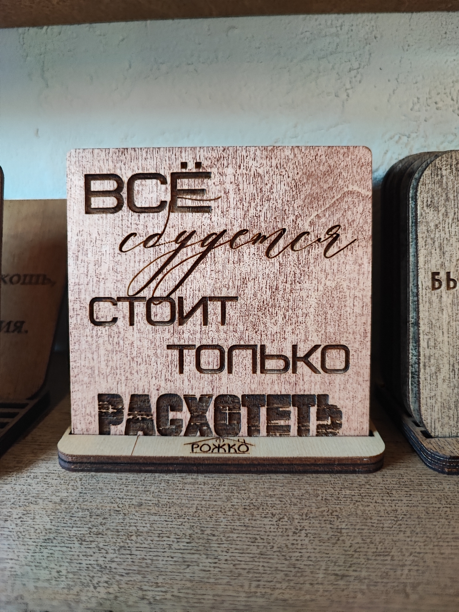 The image displays a wooden plaque with Russian text engraved on it, positioned on a wooden surface, possibly a shelf. The plaque is made of light-colored wood with darker, carved lettering. The text on the main plaque reads: "ВСЁ сбудется стоит только РАСХОТЕТЬ" with smaller text below reading "РОЖКО". The overall aesthetic suggests a decorative item, possibly a gift or a piece of home decor. The plaque is slightly angled and sits on a wooden base. To the right of the main plaque, another similar wooden item with engraved text is partially visible. To the left, another wooden object with text is also partially visible. The background is a light-colored, textured wall. The image does not contain any information about the city of Pyatigorsk, Russia, or any people or interactions.