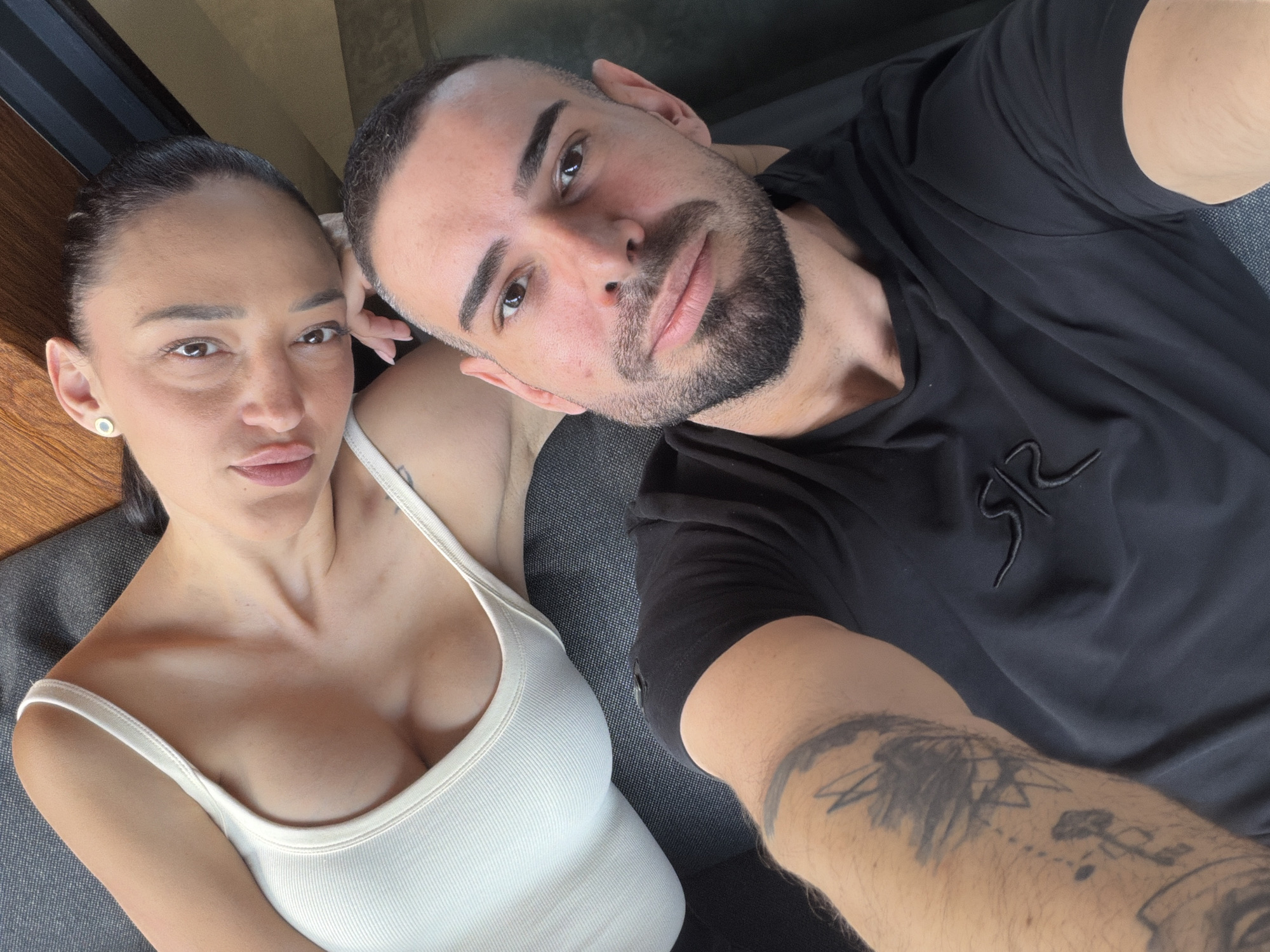 The image is a selfie of a man and a woman. The man has short, dark hair, a beard, and is wearing a black shirt with a logo. He has tattoos on his left arm. The woman has dark hair pulled back, and is wearing a white tank top. They are both looking at the camera. The background is indistinct, but appears to be a grey couch with wooden details behind the woman. There is no indication of the city of Plovdiv, Bulgaria.