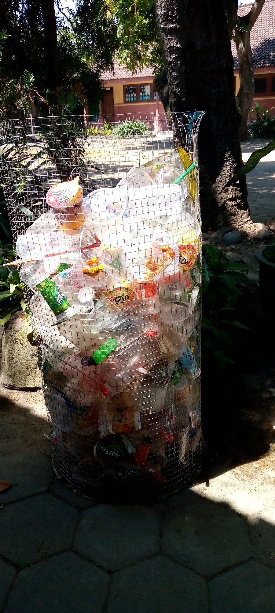 An outdoor scene in Pacing, Indonesia, features a cylindrical wire mesh bin overflowing with plastic waste, primarily bottles and cups. The bin is situated on a paved area with hexagonal tiles. In the background, a building with a tiled roof and a light yellow facade is visible, surrounded by greenery. A large tree trunk is positioned to the right of the bin, casting a shadow. The lighting suggests a sunny day.