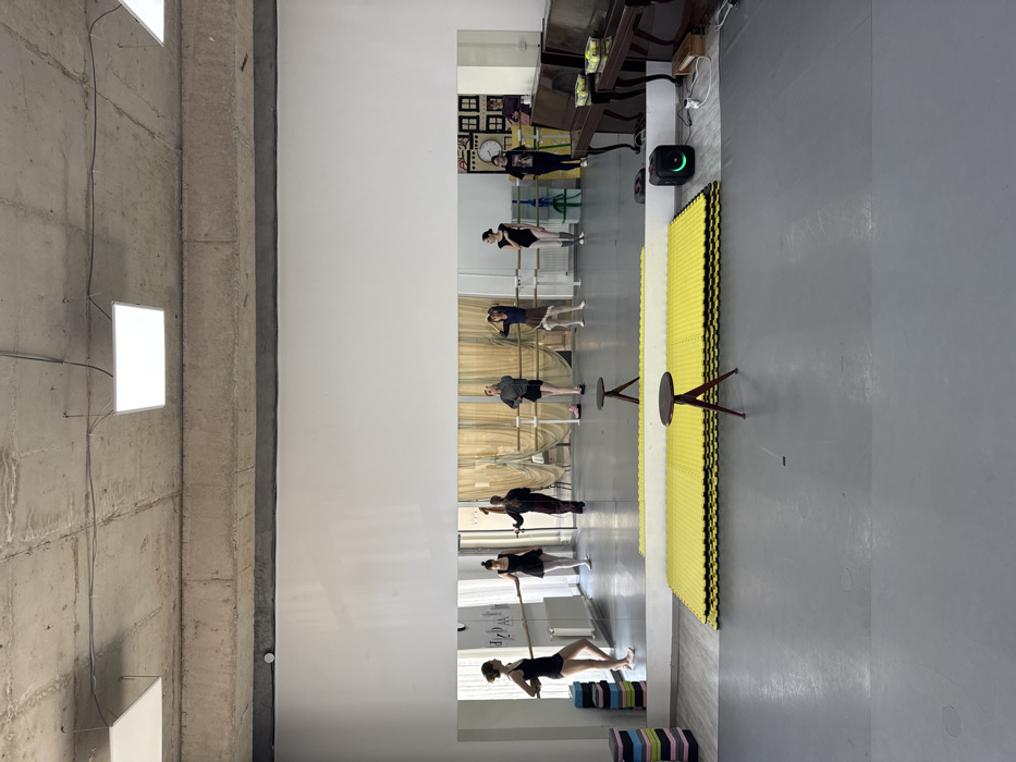 The image displays an indoor dance studio in Tbilisi, Georgia. Seven young women are visible, primarily reflected in a large, floor-to-ceiling mirror that covers the back wall. Most individuals are standing near or holding onto one of two parallel wooden ballet barres mounted at different heights along the mirrored surface. Their attire includes leotards, tights, and athletic clothing.
On the left, a stack of colorful foam exercise blocks is present. In the mid-ground, two small, dark wooden stools with three legs are positioned facing away from the viewer, in front of a rectangular stack of bright yellow interlocking foam mats. To the right, two dark brown upright pianos are side-by-side, topped with several green tennis balls. A black portable speaker with a glowing green light is situated on the floor near the pianos. The studio features a grey floor, white walls, and an exposed concrete ceiling with visible beams and conduits, from which several rectangular LED panel lights are suspended. The reflection also shows a wall decoration resembling framed house shapes and a large clock face beyond the pianos on the right, and a white wall-mounted radiator to the left.