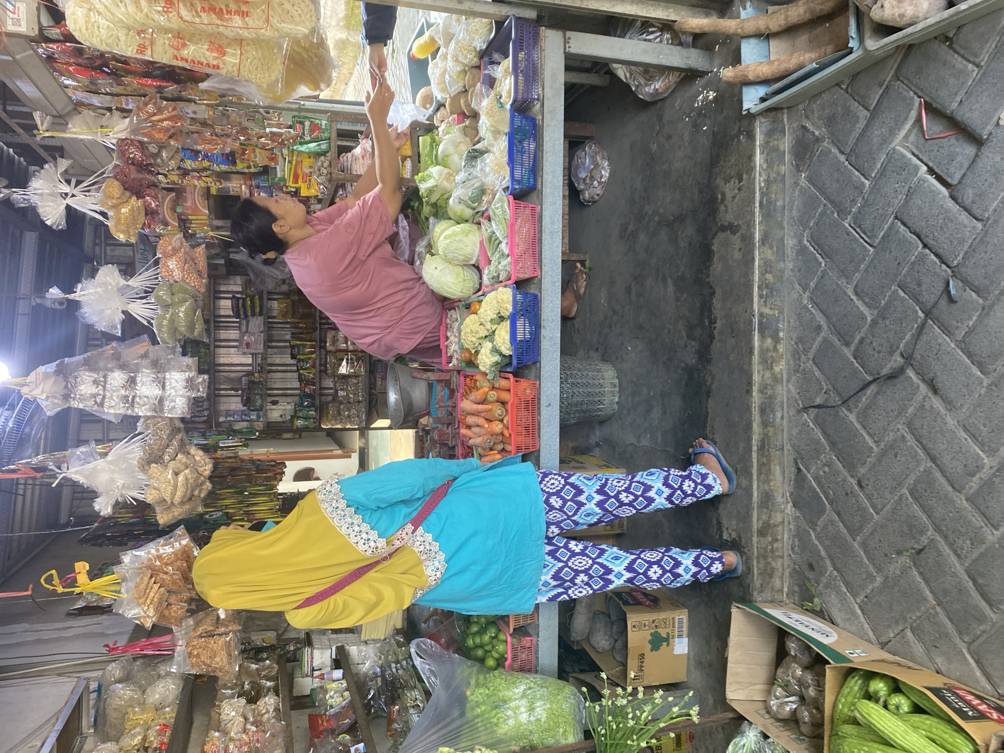 This is a full shot of an outdoor market stall in the city of Kediri, Indonesia, where a vendor is interacting with a customer. The vendor, a woman with dark hair pulled back, wears a pink t-shirt and is leaning over a stall displaying a variety of fresh produce and packaged goods. She is handing something to a customer, whose hand is visible from the elbow down, wearing a dark long-sleeved shirt. The stall is constructed of metal and wood, with shelves above holding bags of snacks and other items. Below, colorful plastic baskets are filled with vegetables such as cauliflower, cabbage, and carrots. A woman wearing a bright yellow hijab and a turquoise top with patterned pants stands facing away from the camera, appearing to be browsing or waiting. In the foreground, boxes of vegetables, including long green gourds and root vegetables, are visible on the ground. The market floor is paved with interlocking grey bricks. The scene is brightly lit, suggesting it is daytime.