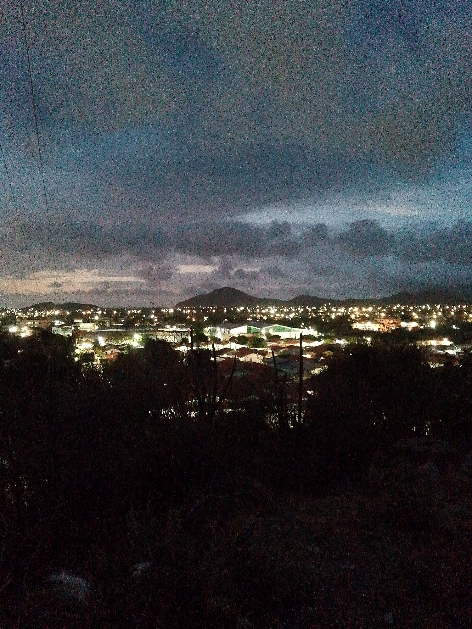 The image depicts a nighttime view of Altagracia, Venezuela, from an elevated vantage point. The cityscape is illuminated by numerous artificial lights, scattered across the urban landscape. In the background, a mountain or hill is visible on the horizon. The sky is overcast with a layer of clouds, giving the scene a dim and muted atmosphere. Foreground vegetation is dark and indistinct, obscuring the lower portion of the image. Power lines can be seen in the top left corner.
