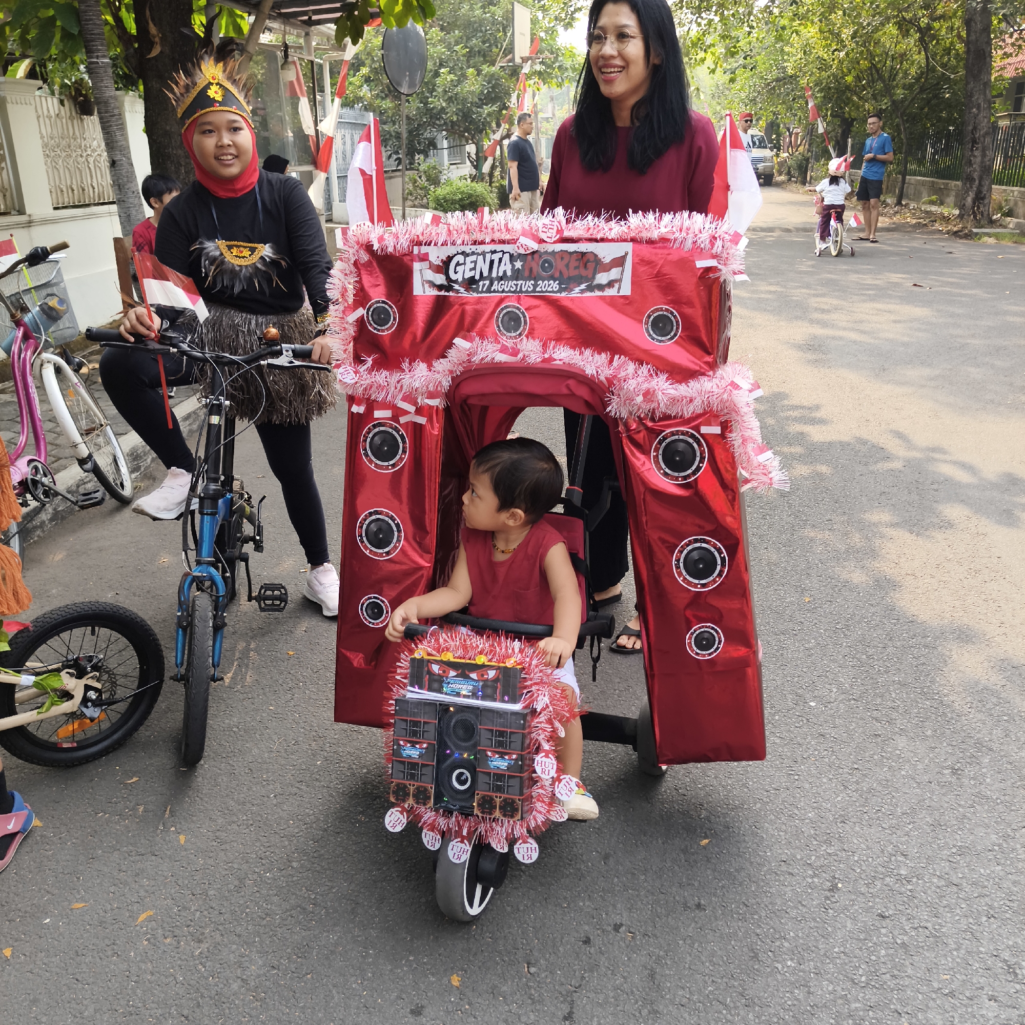 The image depicts an outdoor scene, likely a street in South Jakarta, Indonesia, given the context of an Independence Day celebration (indicated by the Indonesian flags and "17 AGUSTUS 2026" text). A group of people are participating in a parade or event. In the foreground, a child is seated in a large, elaborately decorated red structure resembling a miniature stage or float, which is attached to a small tricycle. The structure is adorned with red and white tinsel and images of speakers. A woman, standing behind the child, is smiling and looking to the right. To the left of the child's tricycle, a young person wearing a red hijab and a decorative headdress, along with a feathered skirt, is seated on a blue bicycle, holding small Indonesian flags. Another bicycle, pink and white, is partially visible to the left. In the background, more people are visible, including a child on a small bicycle and adults standing and observing. The street is paved and lined with trees and buildings. The overall atmosphere suggests a festive community event.