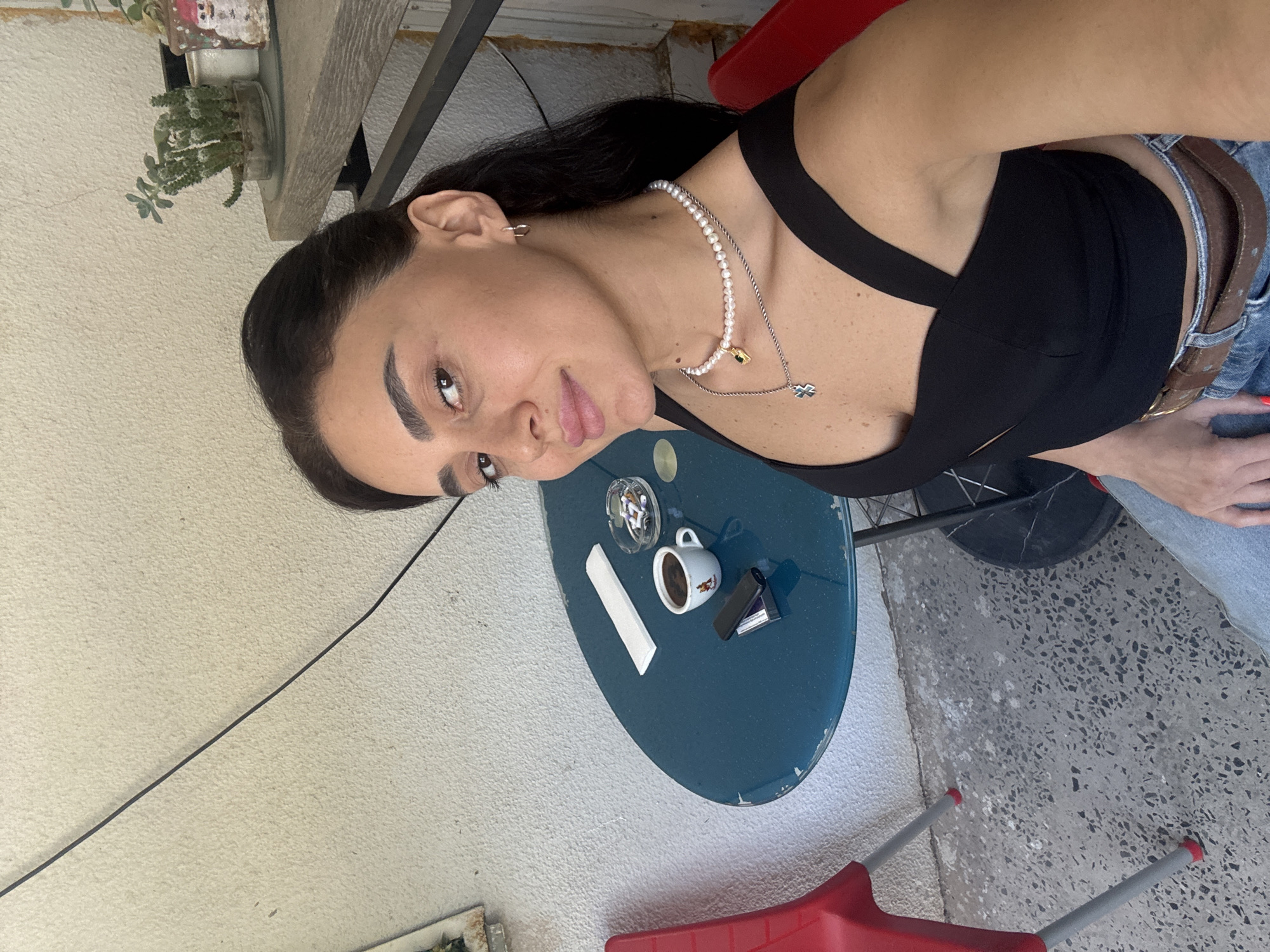 A woman with dark hair and brown eyes is sitting at a round, teal-colored table outdoors. She is wearing a black top and blue jeans, accessorized with two necklaces and small hoop earrings. In front of her on the table is a small cup of coffee, an ashtray with cigarette butts, a lighter, and a napkin. A red chair is visible to her left. The background consists of a textured off-white wall and a shelf with potted plants. The scene appears to be a cafe in Tbilisi, Georgia, given the presence of a cigarette and ash tray, common in outdoor seating areas of cafes.