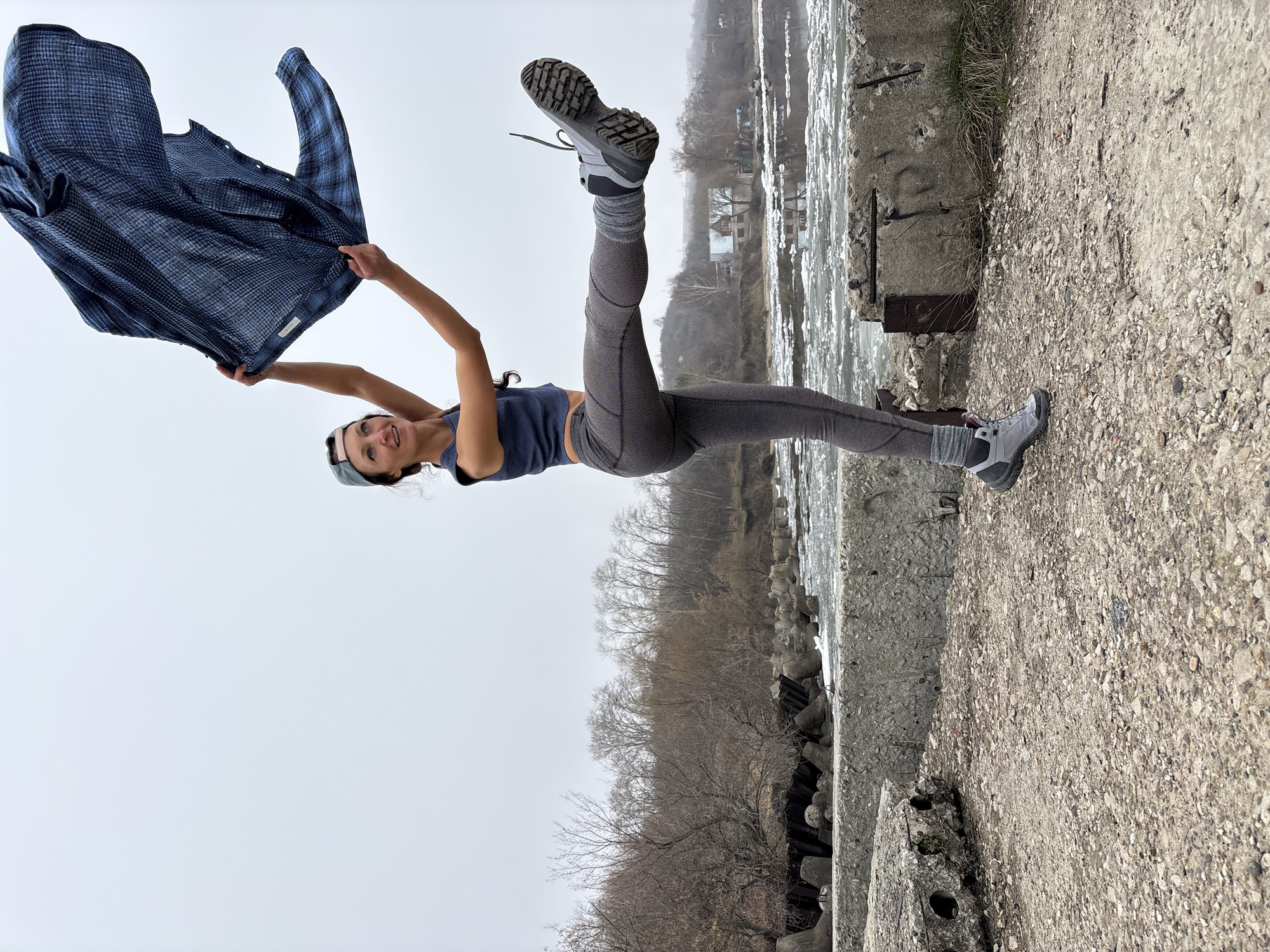 A woman is outdoors, posing in a handstand on a concrete structure, perhaps a pier or embankment, next to a body of water that may be a river or a lake. She is wearing grey leggings, a dark blue top, athletic shoes, and a baseball cap. She is holding a blue plaid shirt overhead. The background includes a body of water with visible ice or turbulent water, trees without leaves, and houses or buildings in the distance. The sky is overcast. It is impossible to ascertain if this photo was taken in Ulyanovsk, Russia.