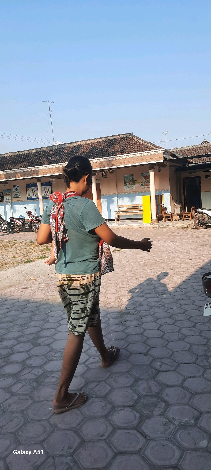 A person, seen from behind and from the side, walks on a hexagonal paved walkway under a clear blue sky. The individual wears a green t-shirt, patterned shorts, and flip-flops. A colorful fabric is wrapped around their torso, supporting a baby also wrapped in fabric, with the baby's legs visible. The person's right arm is extended forward. In the background, a building with a tiled roof and a covered walkway is visible. There are several motorcycles parked to the left and right of the building. The scene appears to be in an outdoor area, possibly a school or community center, in Selopuro, Indonesia, indicated by the "Galaxy A51" watermark in the lower left corner, suggesting the image was taken with that device, and common architecture in the region. A shadow of the person and baby is cast on the ground.