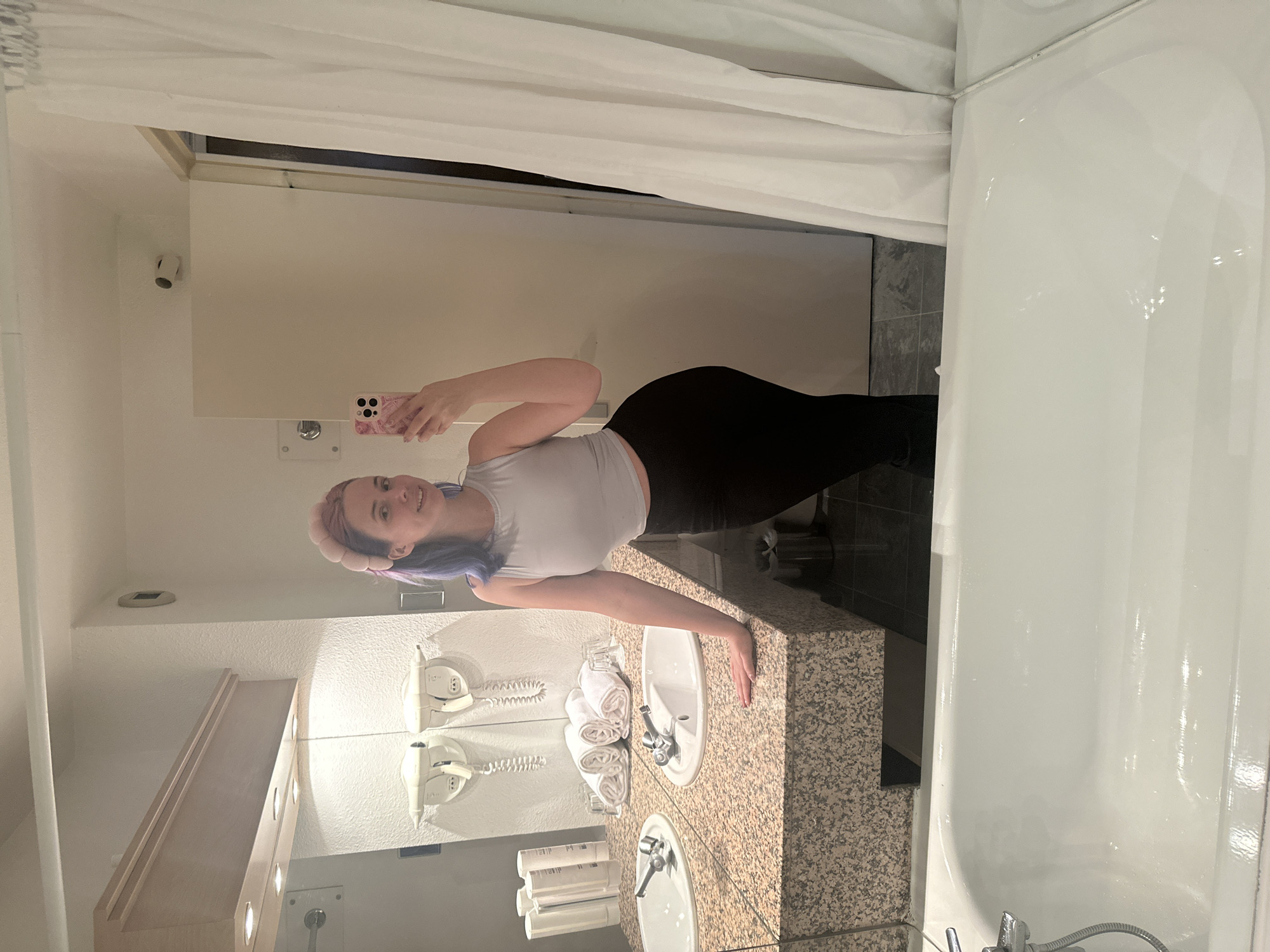 A young woman with purple-streaked hair and a pink headband is taking a selfie in a bathroom. She is wearing a light gray tank top and black leggings. The bathroom has a double vanity with granite countertops, a white bathtub with a shower curtain, and a mirror. The walls are mostly beige. There is a phone on the wall by the mirror and towels and toiletries on the counter. It is not possible to determine the exact location in Ramonville-Saint-Agne, France based on the image alone.