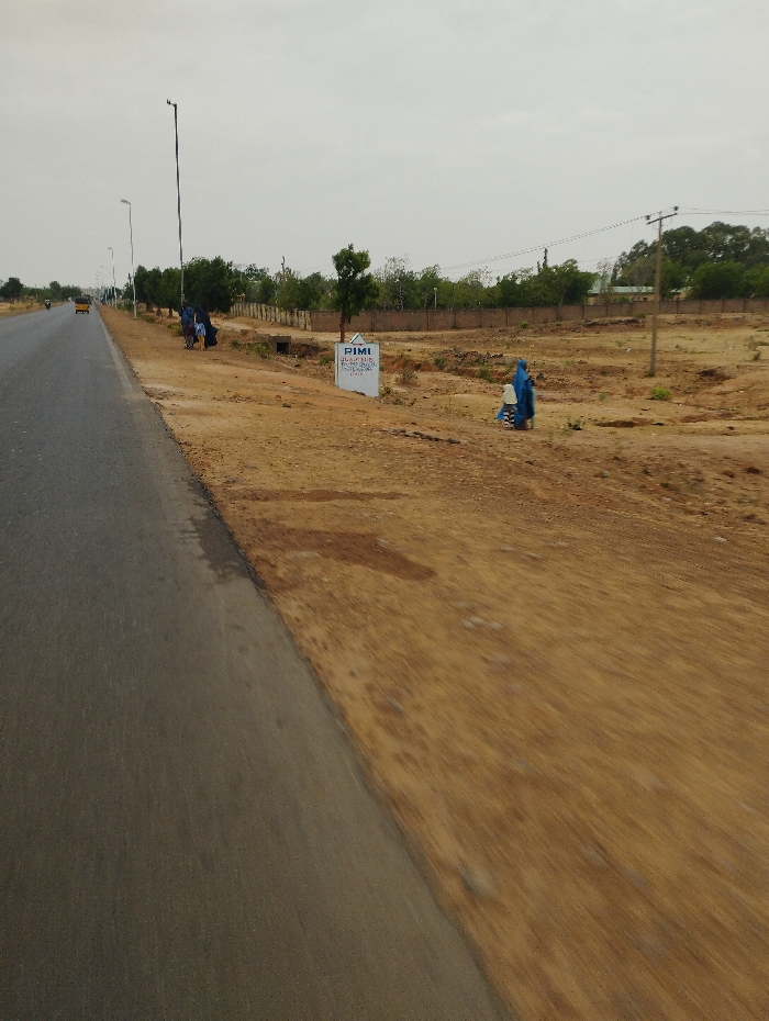 The image shows a paved road in Gabukka, Nigeria, located at coordinates (latitude: 10.263, longitude: 11.139). The road runs from the foreground towards the horizon. There are a few vehicles in the distance on the road. To the right of the road, there is an unpaved area with dirt and dry grass. A sign with the words "PIMI" on it is visible. Three people are walking on the shoulder of the road. Two of them are wearing blue clothing, and they appear to be interacting with each other. The third person is obscured by the other two. The background consists of vegetation, trees, and utility poles. The sky is overcast and the weather appears to be dry. The overall scene is one of a rural landscape with sparse vegetation and few people present.