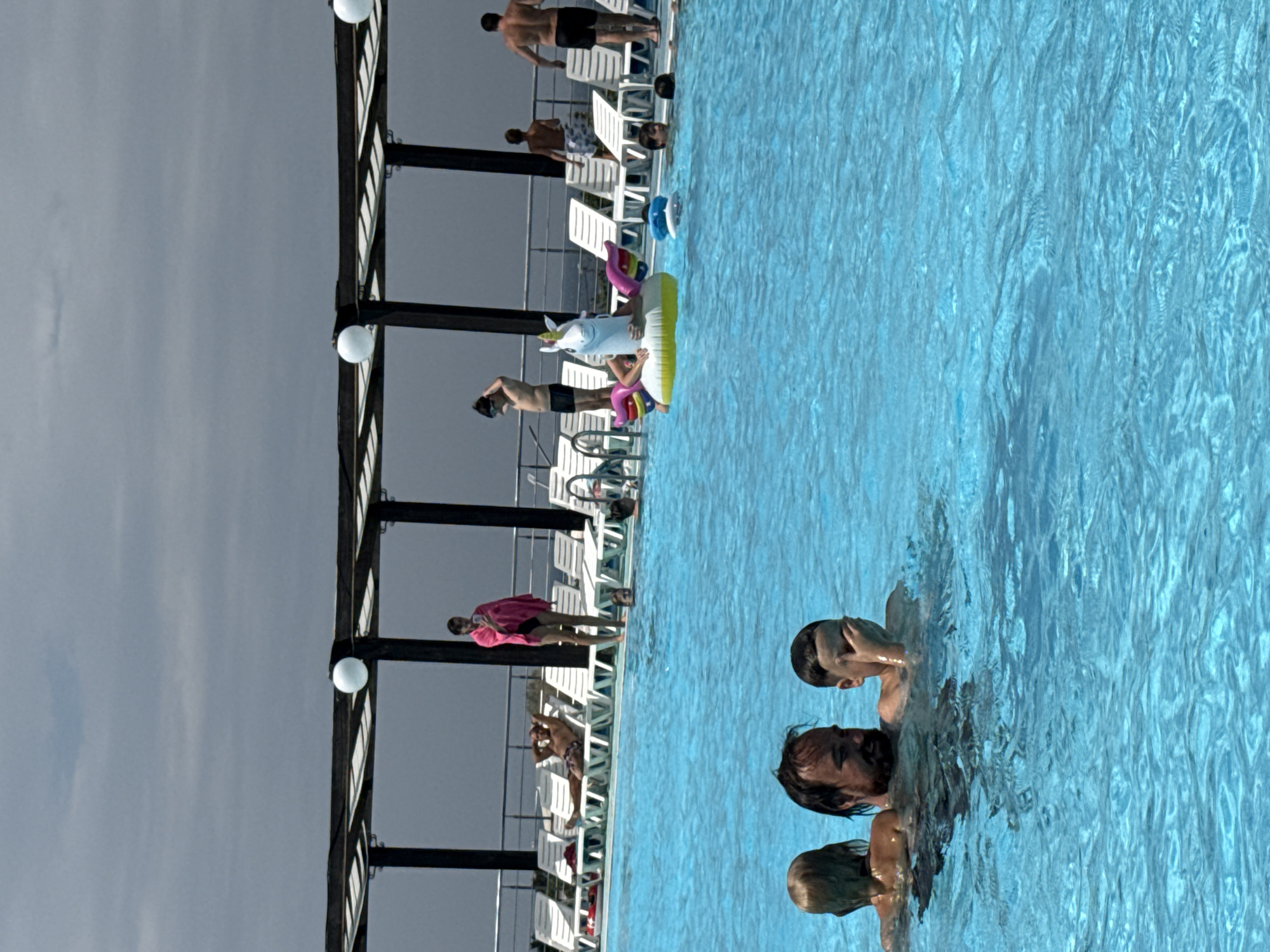 The image depicts a crowded outdoor swimming pool area on a cloudy day. In the foreground, three individuals are partially submerged in the bright blue water. To the left, a blonde-haired person is visible from the shoulders up. In the center, a man with a beard is looking forward, with his head and shoulders above water. To the right of the man, a young person with dark hair is also in the water, covering their eyes with their hand.
In the middle ground of the pool, several people are engaged in various activities. A person is floating on an inflatable unicorn. Another individual is partially visible with a blue flotation device. More people are swimming or wading further back in the pool.
In the background, a paved area with rows of white lounge chairs is visible. Above the lounge chairs, a dark wooden pergola structure with white roof panels extends across the width of the image. Several white spherical lights are attached to the pergola. Beyond the pool area, a metal railing and a slightly hazy sky suggest an open outdoor setting, possibly near a coast or body of water. The overall scene is a lively recreational environment. The presence of the pergola, lounge chairs, and the expansive pool suggests a resort or a public pool facility. Given the specific location of Sevastopol, Ukraine, this could be a popular leisure spot within the city.