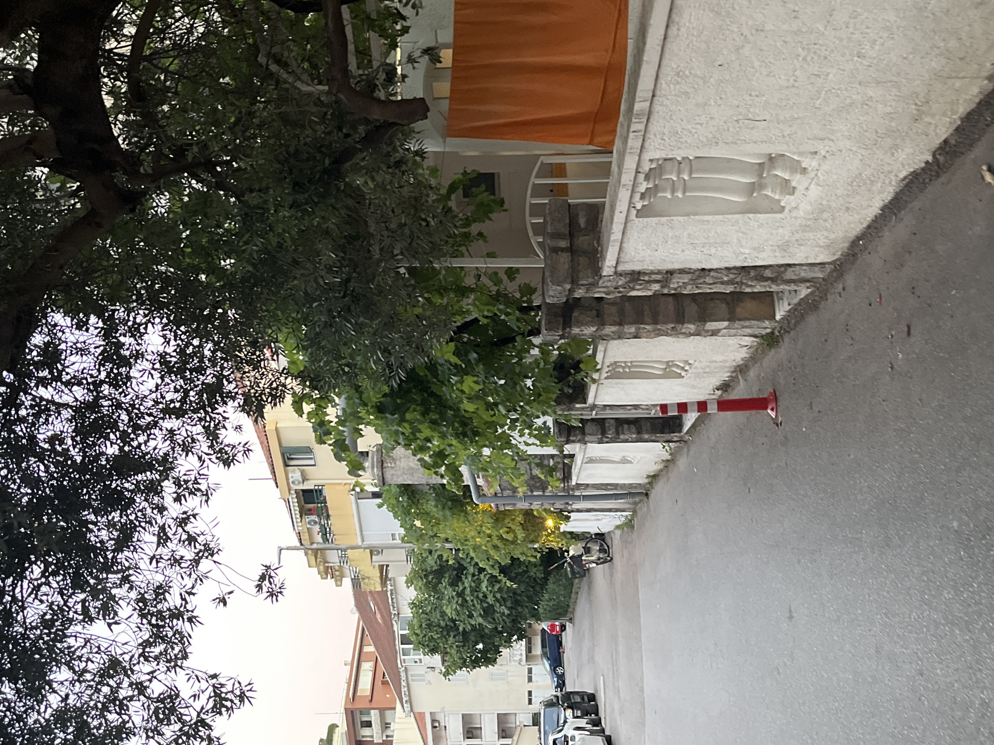 The image depicts a quiet street in Budva, Montenegro, with a low tree canopy overhead casting dappled shadows on the asphalt road. On the right side of the street, a white stucco wall with decorative balustrades separates the road from a property. An orange cloth is draped over what appears to be a railing or clothesline on the right side of the frame. A red and white traffic cone is placed on the road near the wall. Beyond the wall, the facade of a building with white railings is visible. Further down the street, several cars are parked on the left side, including a black SUV and a blue sedan. A bicycle is leaning against a wall on the left side of the road, near some lush green trees. The buildings in the background appear to be residential structures with balconies and air conditioning units. The sky is overcast, suggesting it might be early morning or late afternoon.