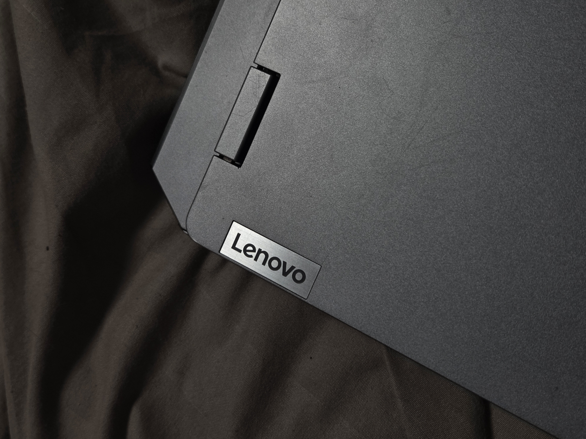 The image shows a gray Lenovo laptop resting on a brown fabric surface. The laptop is positioned at an angle, with the Lenovo logo visible on the lower left corner of the laptop. A rectangular notch or hinge is visible on the top left. The background is out of focus, revealing the texture of the brown fabric. The location is Unknown, Unknown.