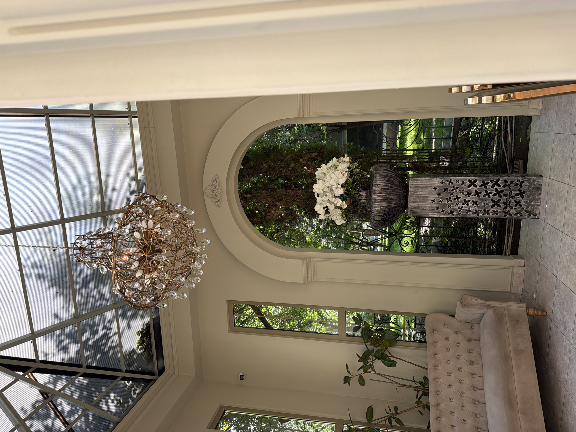 The photograph presents an indoor setting that transitions into an outdoor garden or courtyard. The interior space features a combination of architectural styles including arched openings, rectangular windows, and a glass-paned ceiling, allowing for natural light to penetrate the room. A chandelier with gold-toned metalwork and clear crystals hangs prominently in the left of the frame.
In the background, a large urn filled with white flowers is placed in front of a wrought-iron fence, creating a focal point. A gray, ornate pedestal sits beside the urn. The exterior space is filled with greenery, suggesting a well-maintained garden.
To the right, a tufted sofa in a muted beige color is partially visible, indicating a seating area within the room. A potted plant stands near the sofa, adding a natural element to the interior decor.
The overall impression is that of an upscale, well-designed space blending indoor and outdoor elements. There is no discernible information that suggests the location is Bishkek City, Kyrgyzstan. Therefore, the determination of the photograph originating from this area cannot be confirmed.