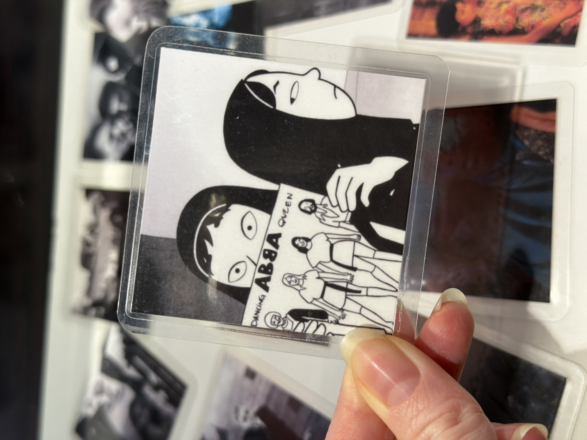 A person's hand, with well-groomed nails, holds up a square-shaped item, likely a magnet or a laminated print, encased in clear plastic. The item displays a black and white graphic-novel style illustration featuring two stylized cartoon figures with dark hair. One figure is depicted in profile, looking upwards, while the other faces forward with wide, staring eyes. Between these figures, a small rectangular object resembling an album cover or book is shown. Clearly visible text on this object reads "DANCING ABBA QUEEN," with "ABBA" prominently displayed, stylized in a way that the 'B's resemble the number '8'.
The background, largely out of focus, reveals several other similar square or rectangular items, suggesting a collection displayed on a surface, possibly a refrigerator or magnetic board. Most background items appear to be black and white photographs or prints, though one shows hints of warm colors like red and orange. The lighting is soft and even, highlighting the details of the held item and the hand. The scene is set indoors, likely in a home, and the image was taken in Khimki, Russia.