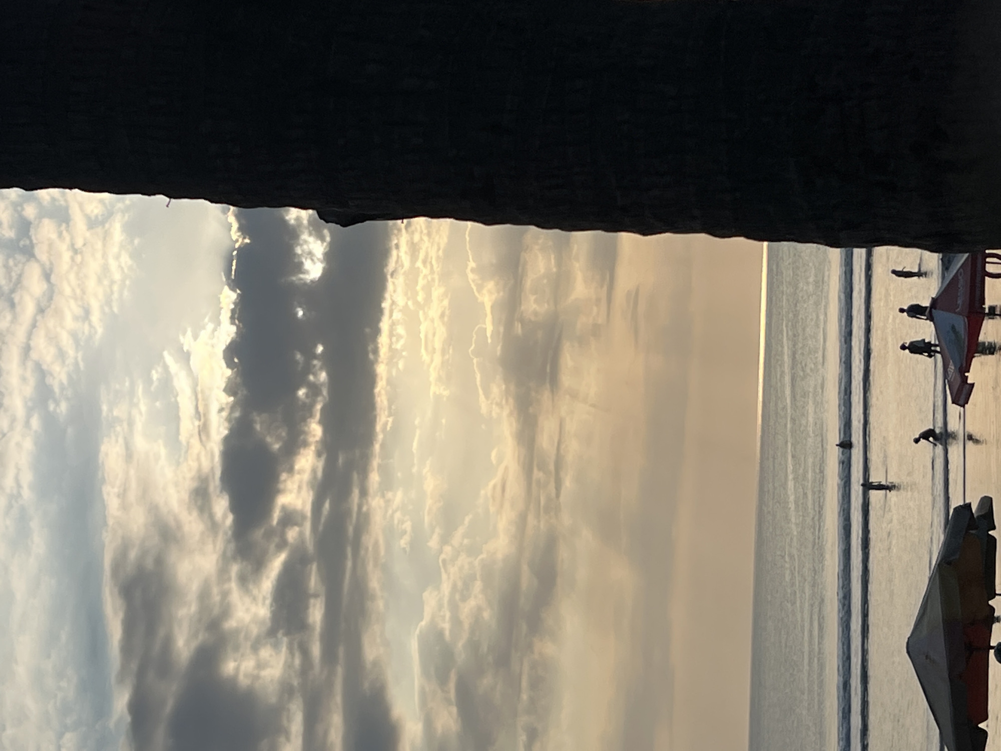 The image depicts a beach scene in Kuta, Indonesia, during what appears to be sunset or sunrise. A large, dark silhouette of a tree trunk dominates the right side of the frame. To the left, a serene ocean with gentle waves stretches towards the horizon, where the sky is filled with a soft, warm glow. The sky is a canvas of dynamic clouds, with rays of sunlight piercing through them, creating dramatic light effects. Several people are visible in the shallow water, some appearing to walk or stand, while others are wading. In the foreground, beach umbrellas are partially visible, suggesting a recreational area. The overall atmosphere is peaceful and tranquil, capturing the beauty of a tropical coastal environment.