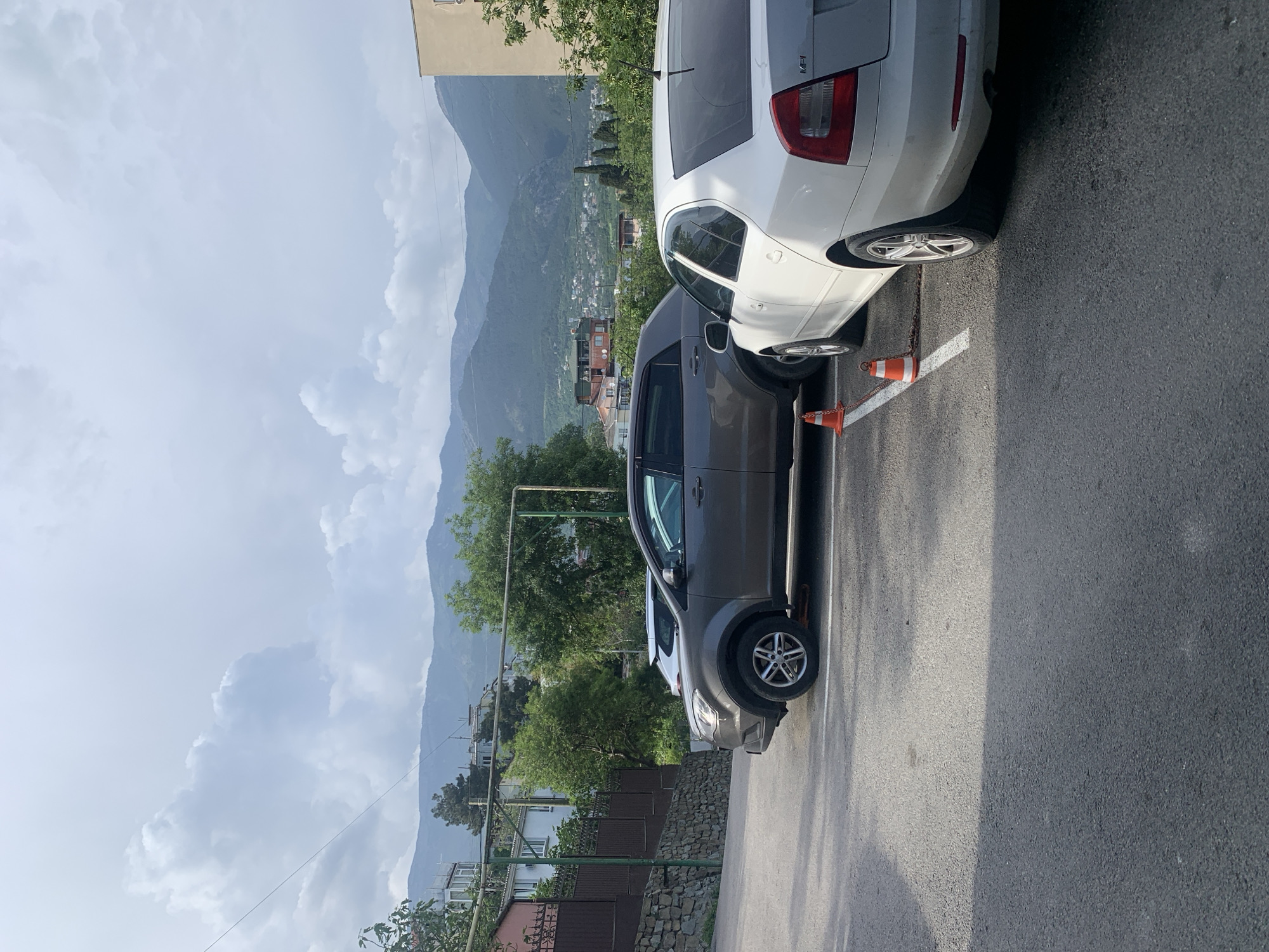 The image shows a street scene in an urban or suburban area. In the foreground, there are two cars parked adjacent to each other: a dark-colored sedan or hatchback and a white sedan. Orange traffic cones are positioned between the two cars, possibly indicating a restricted or hazardous area. The road surface appears to be asphalt.
In the background, there's a view of a mountainous landscape, suggesting the location is near a mountain range. Buildings and vegetation are visible further in the background, indicating a town or city nestled in the valley. The sky is overcast with clouds.