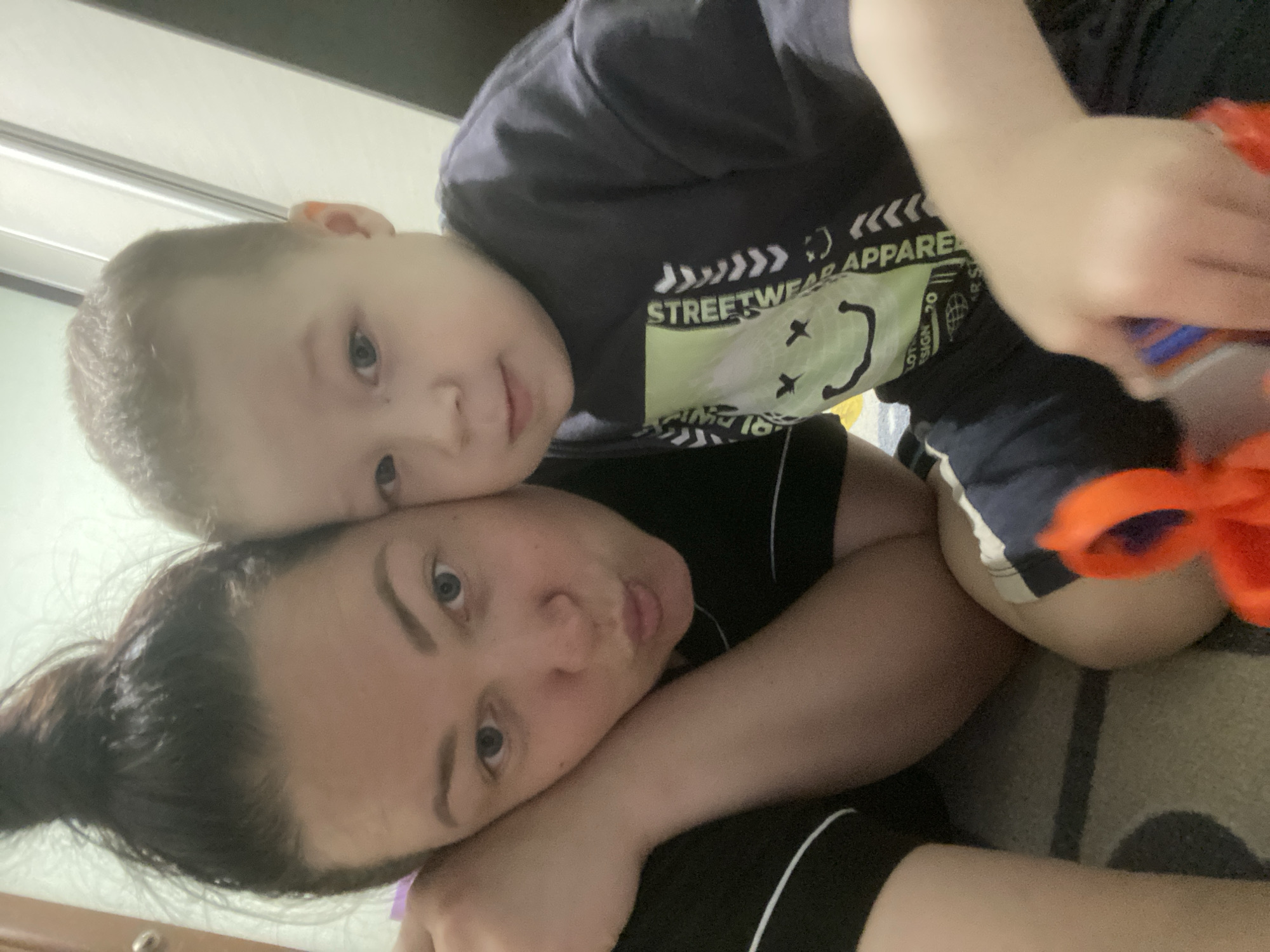 A woman and a young child are lying down together. The woman is lying on her side with her head resting on a surface. Her dark hair is pulled back into a ponytail. The child is lying on top of her, face close to hers, wearing a dark blue t-shirt. The shirt has a graphic design with "STREETWEAR APPAREL" printed on it. The child is also wearing dark shorts. The background is a blurry interior with a closed door and a carpeted floor. The city of Sochi, Russia, cannot be determined from this image.