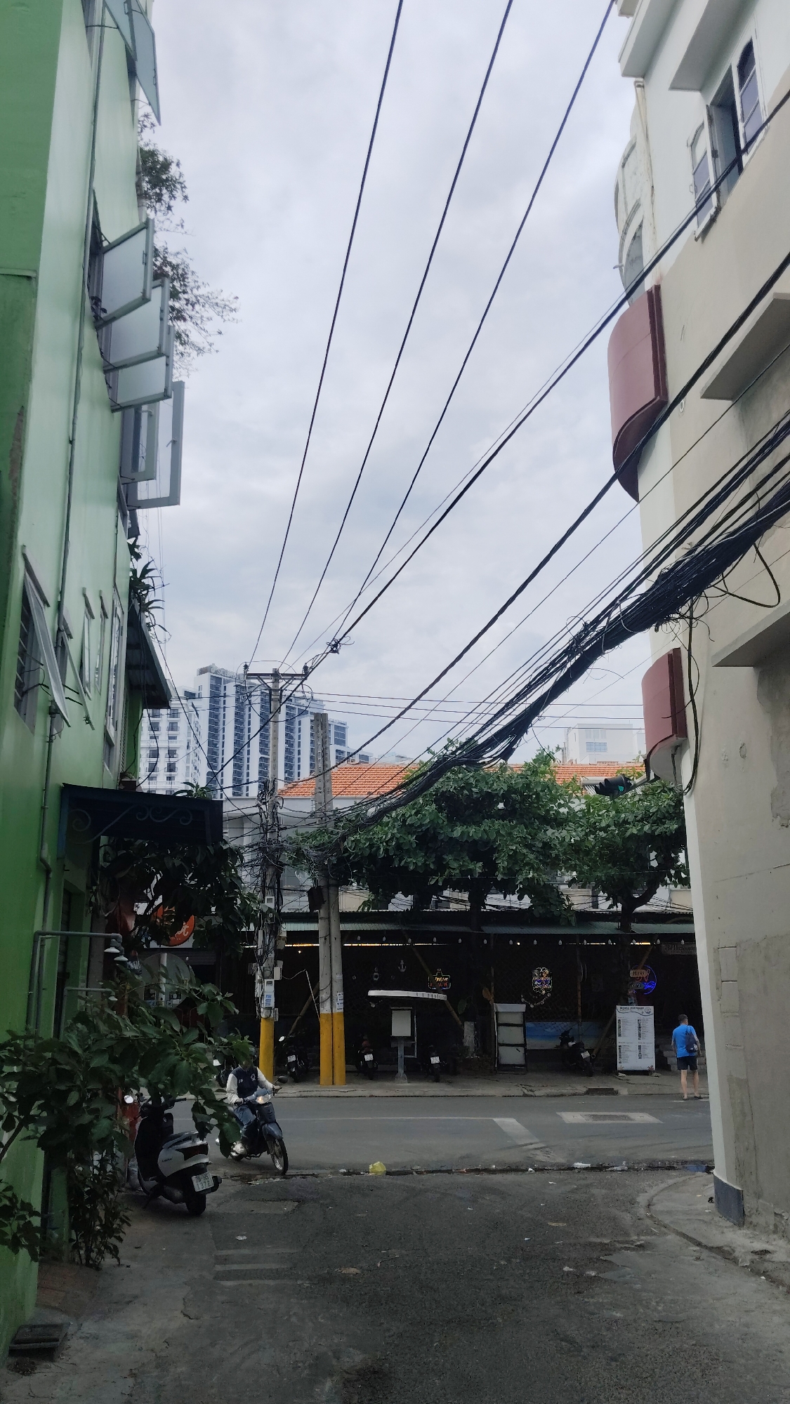 The image shows a narrow street scene in Bac Nha Trang Ward, Vietnam. The street is flanked by buildings, one painted light green and the other painted off-white. Numerous power lines crisscross overhead. A line of trees runs along the center of the road. There are motorbikes parked near a bar or restaurant that has nautical decor such as anchors, which sits under the trees. A man in a blue shirt is walking to the right of the image. Across the street, in the distance, there is a view of tall buildings. The sky is overcast.