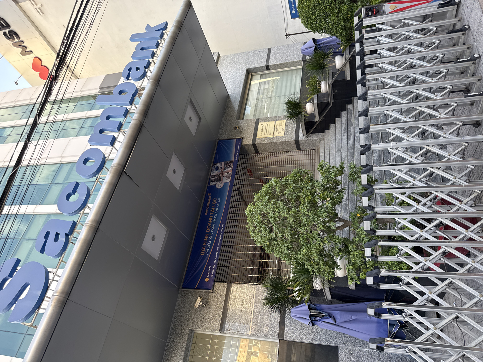 The exterior of a Sacombank branch building is pictured. The bank's name is displayed in large blue letters on the facade. In front of the entrance, a metal accordion gate is closed and extended, blocking access to the building. Two closed blue umbrellas stand to the left of the entrance stairs. Several potted plants are placed on the steps leading up to the bank. A banner advertising "GÓI KINH DOANH TÀI LỘC" (Prosperity Business Package) is visible above the entrance, along with a security camera. The building appears to be located in the city of Nha Trang, Vietnam, as indicated by the signage and architectural style common in Vietnamese cities. The scene suggests the bank is closed, possibly outside of business hours.