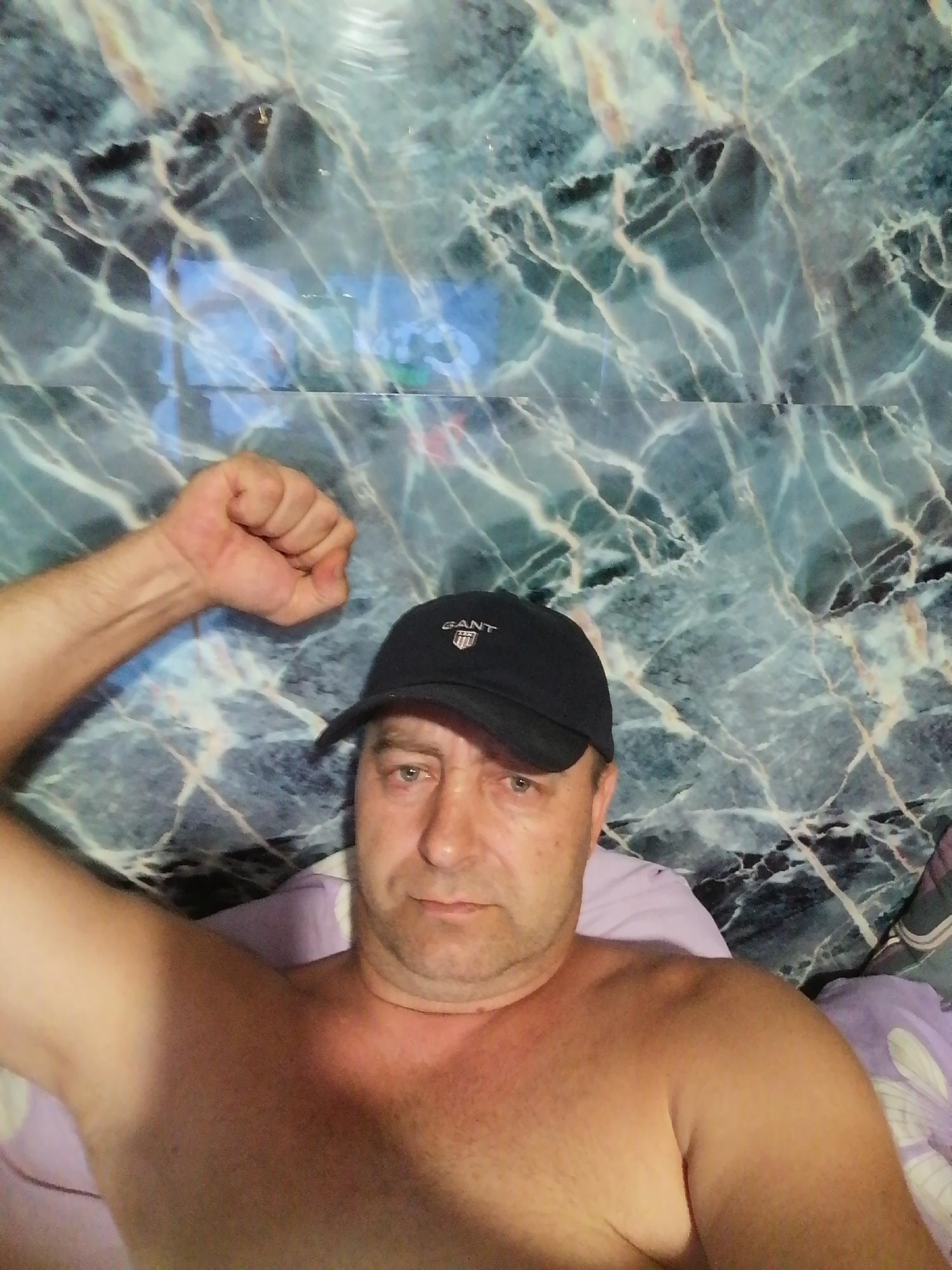 A man wearing a black baseball cap with a "GANT" logo is lying down and looking at the camera. He is shirtless, and his right arm is raised with his fist clenched. The background features a marbled wall with green and blue hues. The image appears to be a selfie.