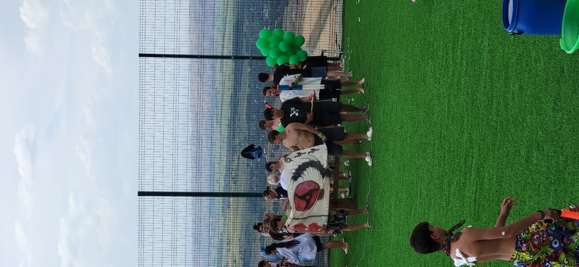 The image depicts an outdoor gathering of young people on a grassy area, likely a recreational space. The setting appears to be elevated, with a wire fence in the background overlooking a valley with scattered buildings and hills under a cloudy sky.
In the foreground, a young boy is visible from the back, wearing colorful shorts adorned with cartoon monsters. He has a bandana around his neck and some white material or paint on his back. He is holding an orange water gun.
In the middle ground, a group of teenagers and young adults are standing, some holding a large white banner with a red symbol resembling an eye and black swirling patterns. Several individuals are shirtless, suggesting warm weather. Some are holding water guns. To the right of the group, a cluster of green balloons is visible.
The overall atmosphere suggests a casual event, possibly a water fight or a playful gathering, taking place in a scenic outdoor location in Сырымбет, Kazakhstan.
