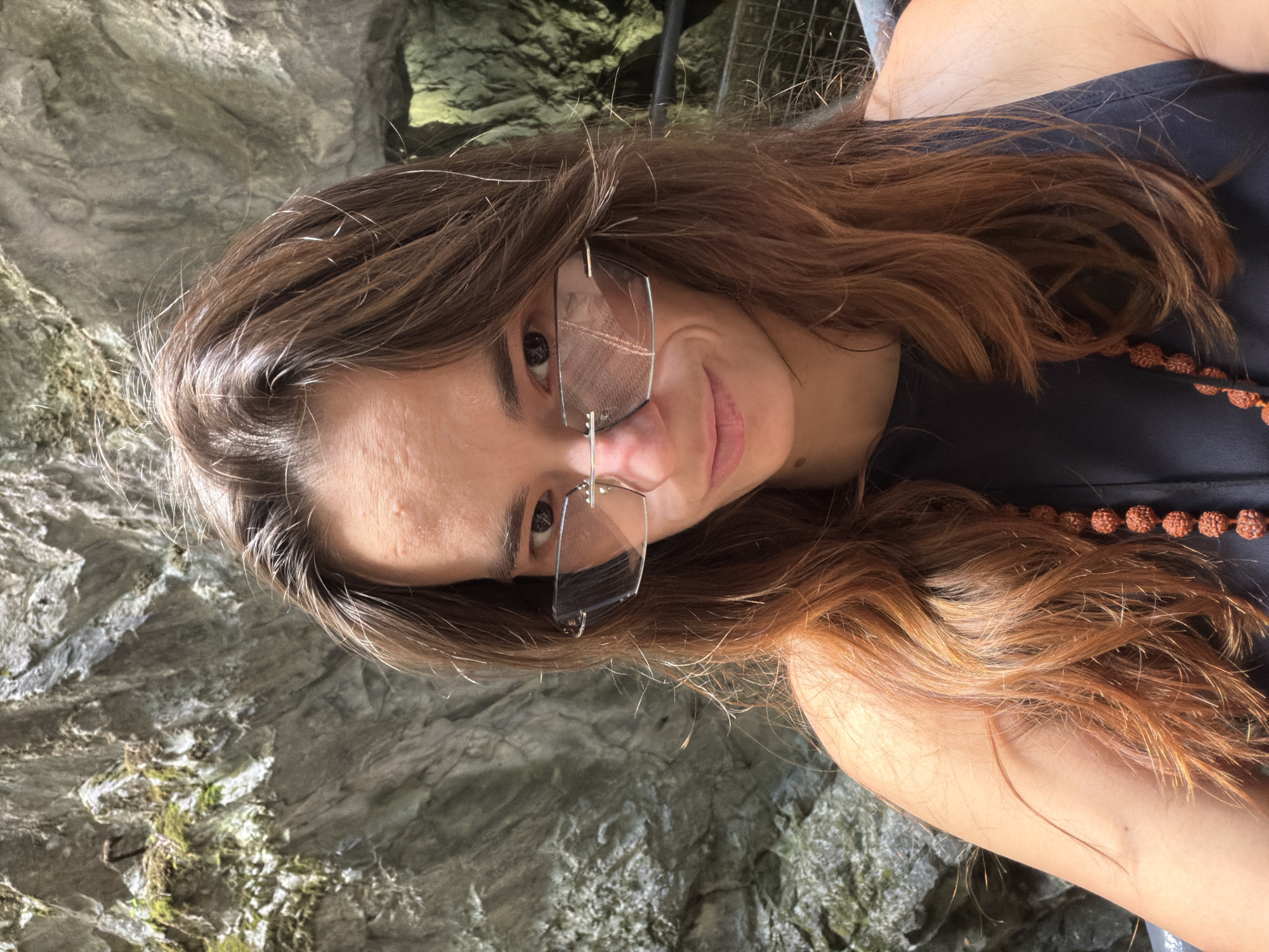 A young woman with long, auburn hair is taking a selfie in a narrow gorge. She is wearing sunglasses with a silver frame and a dark top, accessorized with a beaded necklace. The background is a rough, grey rock face, characteristic of the Ruskeala Mountain Park, a former marble quarry in Sortavala, Russia, known for its picturesque gorges and clear water. The lighting suggests it is daytime, with sunlight filtering into the shaded gorge.