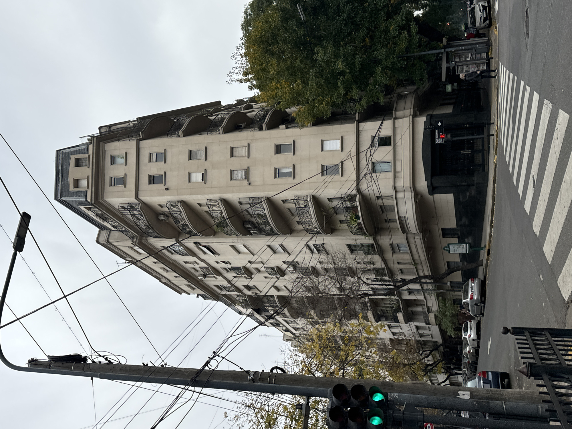 The image captures a street scene in Buenos Aires, Argentina. A tall, light-colored building with multiple stories dominates the view. The building features wrought iron balconies and a complex roofline with arched details. A large, green tree partially obscures the upper portion of the building.
Below the building, on the street level, several parked cars are visible. A crosswalk with white stripes stretches across the street. A pedestrian is crossing the street and a traffic light displays a green signal.
In the foreground, a utility pole is laden with wires. The sky above is overcast and gray.