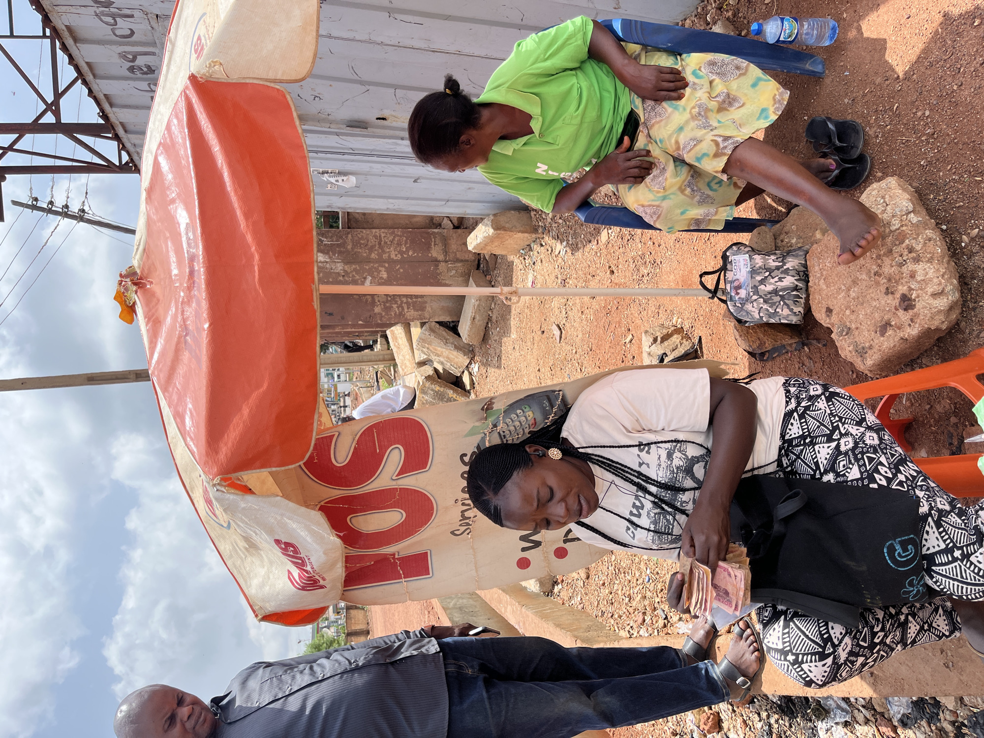 A scene in Gura Top, Nigeria, shows two women sitting outside under a partially visible orange and white advertising umbrella for "SOS White Detergent." One woman wears a green top and yellow skirt and is barefoot on a rock. Near her are a pair of black sandals, a camouflage bag, and a bottle of water. The other woman, wearing a white t-shirt and black and white patterned pants, sits on an orange plastic chair, counting a stack of money. A black tote bag sits on her lap. A man in a gray shirt and blue jeans stands near her. The ground is dirt and gravel, with scattered debris. A metal fence is visible behind the women. Utility poles and wires are partially visible in the background.