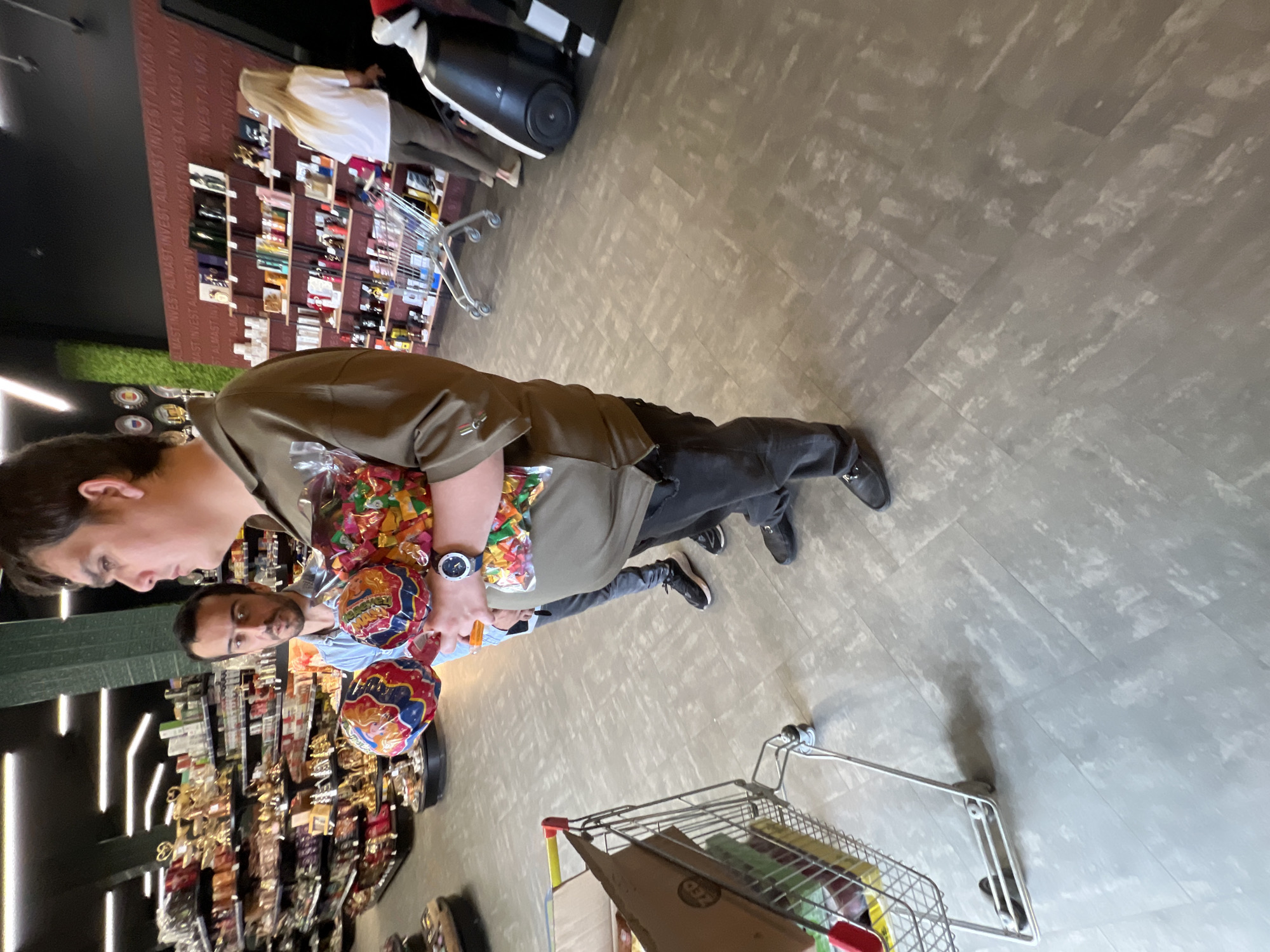 This image captures an indoor scene within a commercial establishment, likely a supermarket, department store, or gift shop, located in Krasnodar, Russia. The photograph is oriented sideways.
In the foreground, a man in a brown/khaki shirt, dark pants, and a dark wristwatch on his left wrist holds several colorful, transparent bags filled with what appear to be candies or small toys. His face is visible in profile, looking downwards. Behind him, a second man with a beard, wearing a light blue shirt, is partially visible, also looking towards the side.
Further in the background, a woman with long blonde hair, dressed in a white top and dark pants, is seen near a section of shelves. The shelves lining the walls display numerous products, suggesting items such as cosmetics, perfumes, or small gifts. One shopping cart, partially filled with items including a cardboard box marked "ZED," is in the lower right of the frame, and another is visible in the mid-background. The floor is a light gray with a subtle patterned design.
A prominent feature in the background is a reddish-brown wall section adorned with the repeated text "ALMAST INVEST," which provides a strong location cue. Variations of this text like "ALVASTING" and "ALMAST INVERT" are also discernible. The lighting is bright and artificial, typical of a retail environment. The individuals appear to be engaged in shopping or browsing. No strong emotions are evident on their faces.