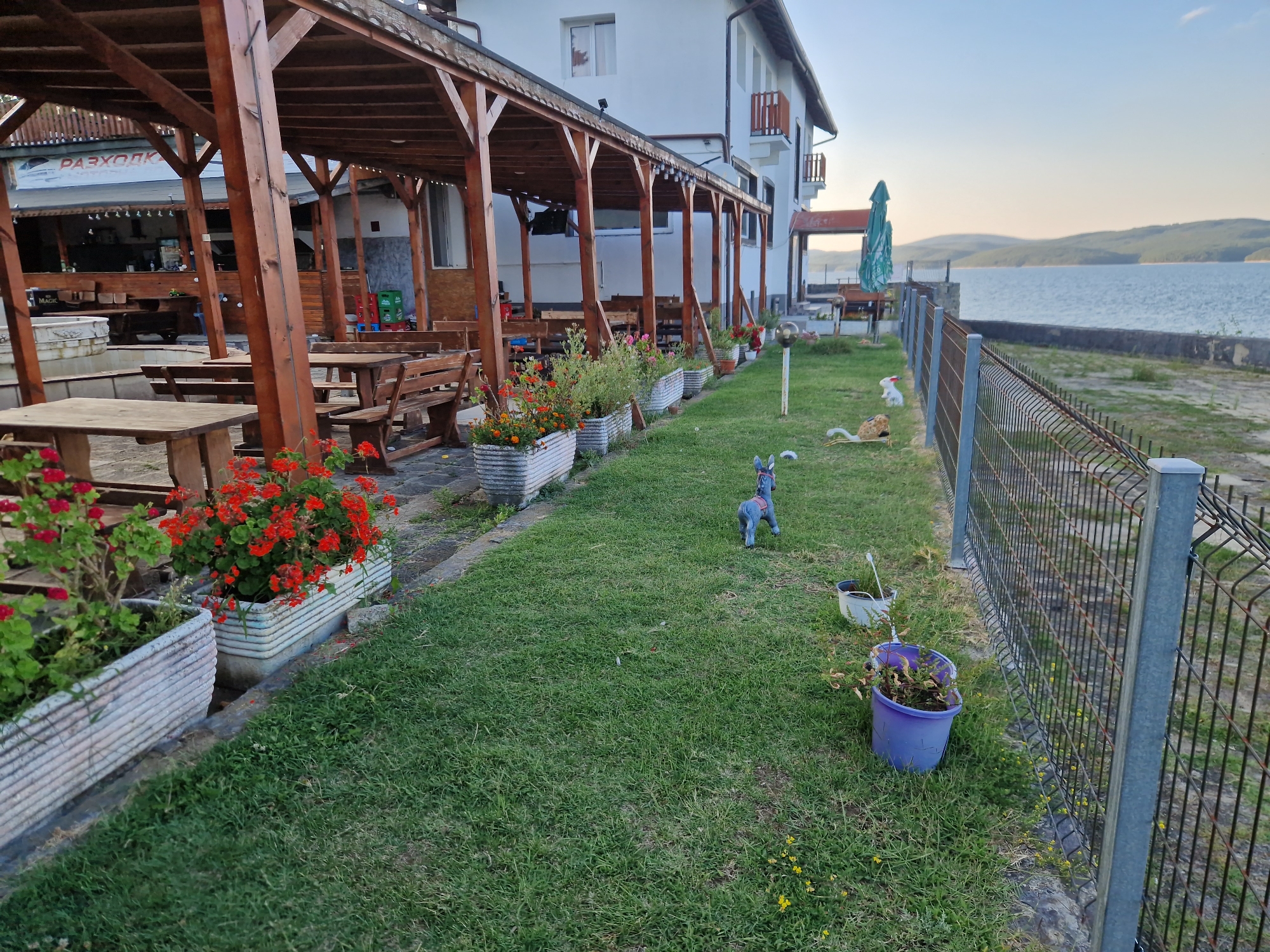 The image depicts an outdoor seating area adjacent to a body of water, likely a lake or reservoir, with a two-story building in the background. The foreground features a grassy lawn with several decorative garden elements, including flower planters with red and pink blooms, a blue donkey figurine, and a white cat figurine. A metal fence runs along the right side of the image, separating the lawn from the water. Wooden picnic tables and benches are visible under a covered patio area to the left. The building appears to be a restaurant or guesthouse, with a sign reading "РАЗХОДКИ" visible on the left side. The overall setting suggests a tranquil, waterside location. The image was taken in the vicinity of Dolni Pasarel, Bulgaria.