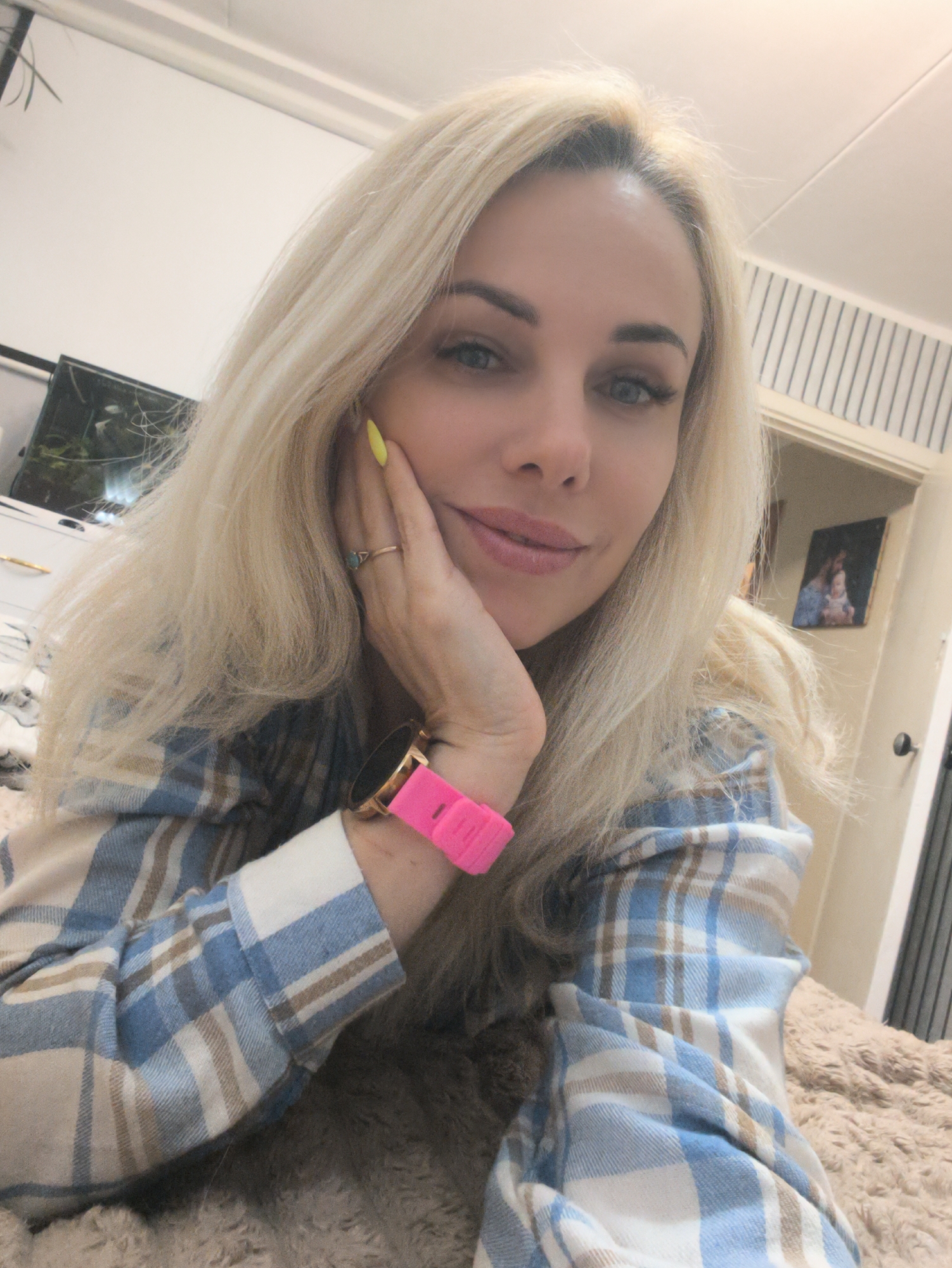 A woman with blond hair and blue eyes is lying down, resting her face on her hand. She is wearing a blue and white plaid shirt and a pink smartwatch. She has a ring on her finger and her nails are painted yellow. In the background, there is a television, a bed, a doorway, and a picture hanging on the wall. There is no indication this picture was taken in the city of Sevastopol, Ukraine.