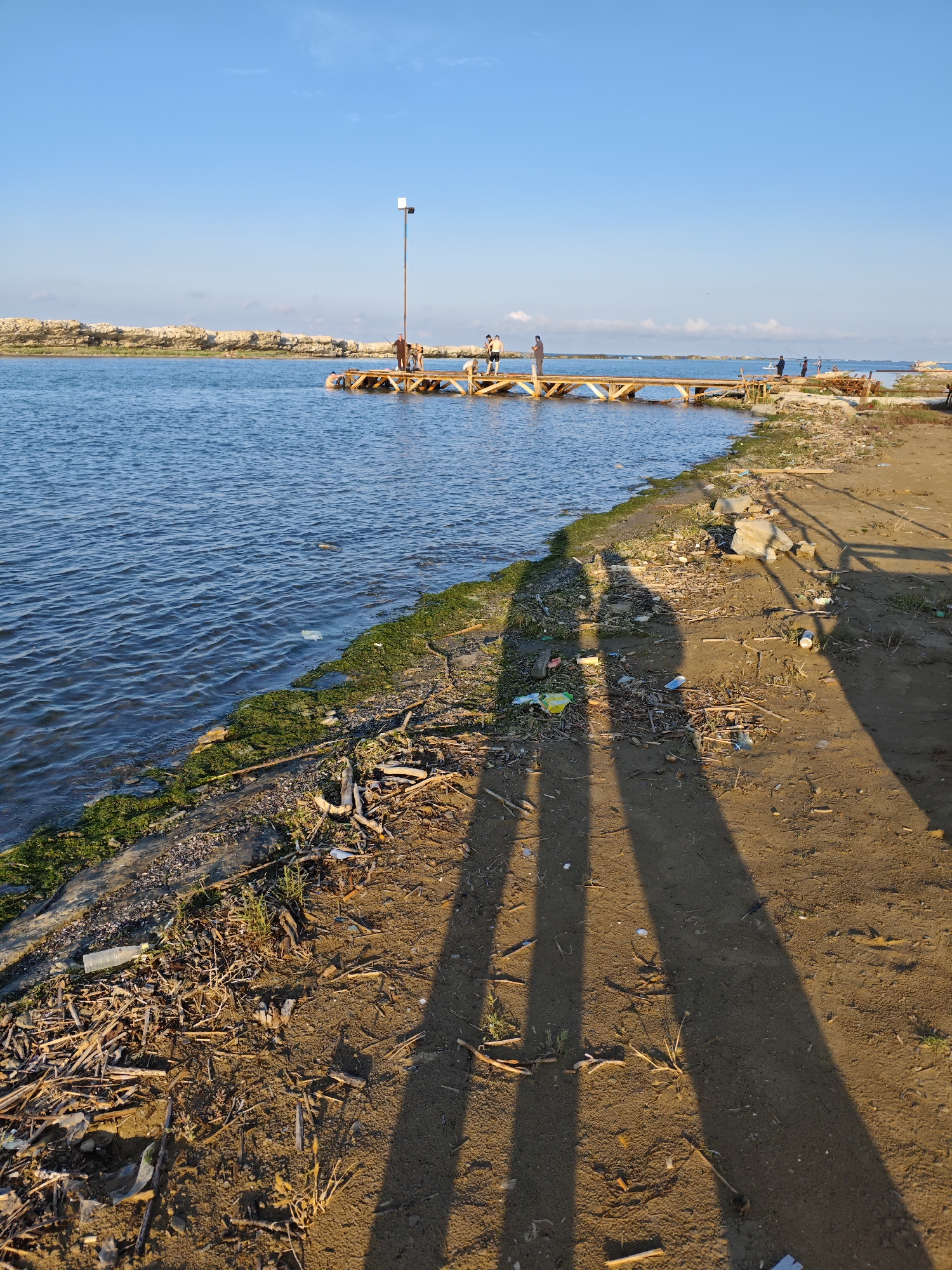 The image depicts a sunny day at a coastal area, likely a beach or embankment, in Izberbash, Russia. In the foreground, the sandy ground is littered with debris, including sticks, leaves, and plastic waste. Two distinct shadows of people are cast across the sand, indicating their presence in the scene, though they are not fully visible.
The middle ground shows a body of blue water, with gentle waves lapping against the shore. A wooden pier extends into the water, with several people visible on it. Some appear to be fishing, with fishing rods in hand, while others are standing or walking. A tall pole with a light fixture stands on the pier. The shoreline is lined with green algae and more debris.
In the background, a rocky formation forms a distant coastline. The sky is a clear blue with a few wispy clouds. The overall impression is a casual, outdoor gathering with elements of both leisure and environmental neglect due to the visible trash.