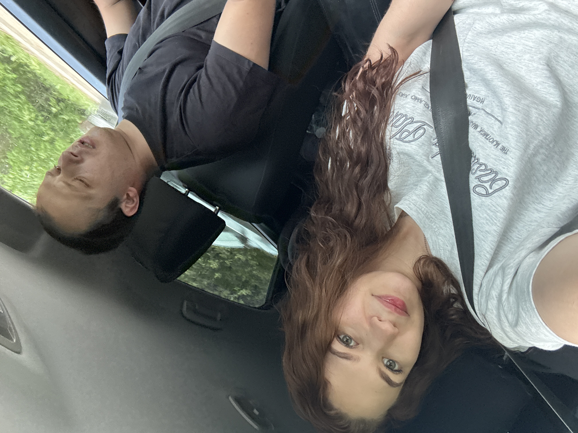Two people are in a car, both wearing seatbelts. The person on the left is a man wearing a black shirt and is reclined with his eyes closed. The person on the right is a woman with long, wavy brown hair wearing a gray t-shirt. Outside the car window, blurred green foliage is visible. The interior of the car is dark. The location of Chaikovsky, Russia, is not discernible from this image.