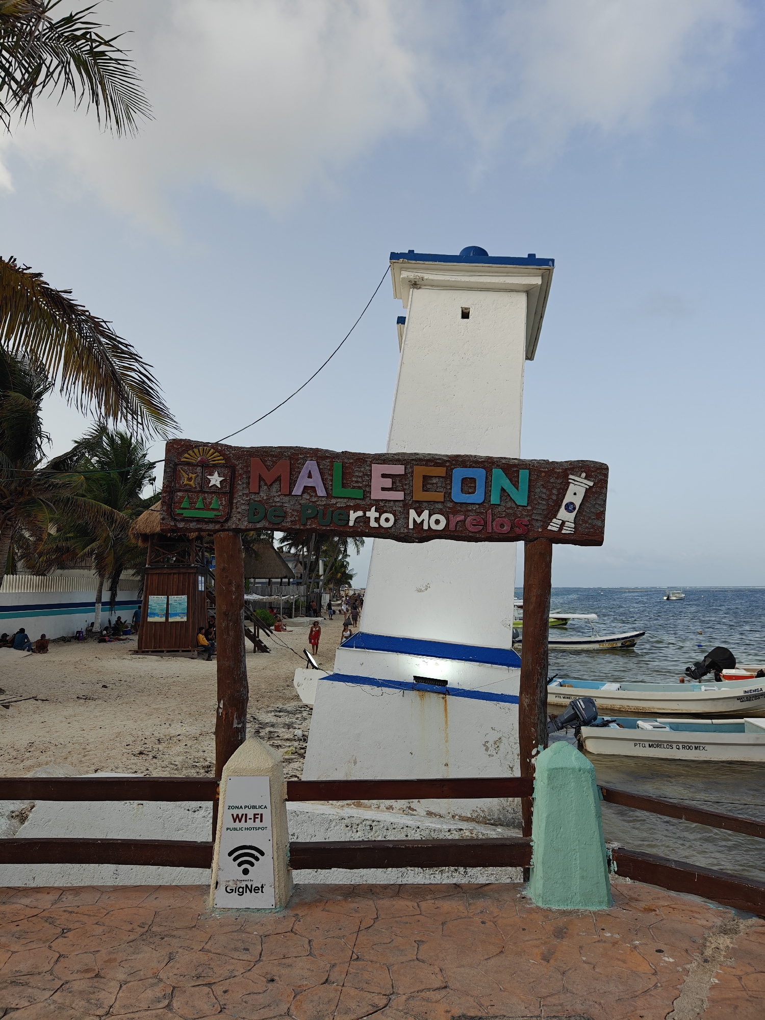 This is a medium shot of the Malecón in Puerto Morelos, Mexico, on a partly cloudy day. The foreground features a paved walkway with an orange-toned, stamped concrete surface. A brown wooden railing runs horizontally across the frame, with a green concrete bollard positioned on the right side of the walkway. To the left of the bollard, a white sign with a black border indicates "ZONA PÚBLICA WI-FI PUBLIC HOTSPOT" with a Wi-Fi symbol and the GigNet logo at the bottom.
A tilted white lighthouse structure with blue accents is a prominent subject. Attached to a wooden post in front of the lighthouse is a colorful wooden sign that reads "MALECÓN De Puerto Morelos". The sign features an emblem with a sunburst, star, and trees on the left, and a depiction of a lighthouse on the right.
Behind the sign and lighthouse, a sandy beach is visible. Several people are scattered across the beach, some sitting and others walking. A small wooden structure with a thatched roof and a blue and white striped wall stands on the beach. Further back, palm trees with fronds overhang the scene.
The water of the Caribbean Sea is visible in the background, with several small motorboats docked or anchored. The sky is a pale blue with scattered white clouds. The overall impression is a relaxed, coastal resort atmosphere.