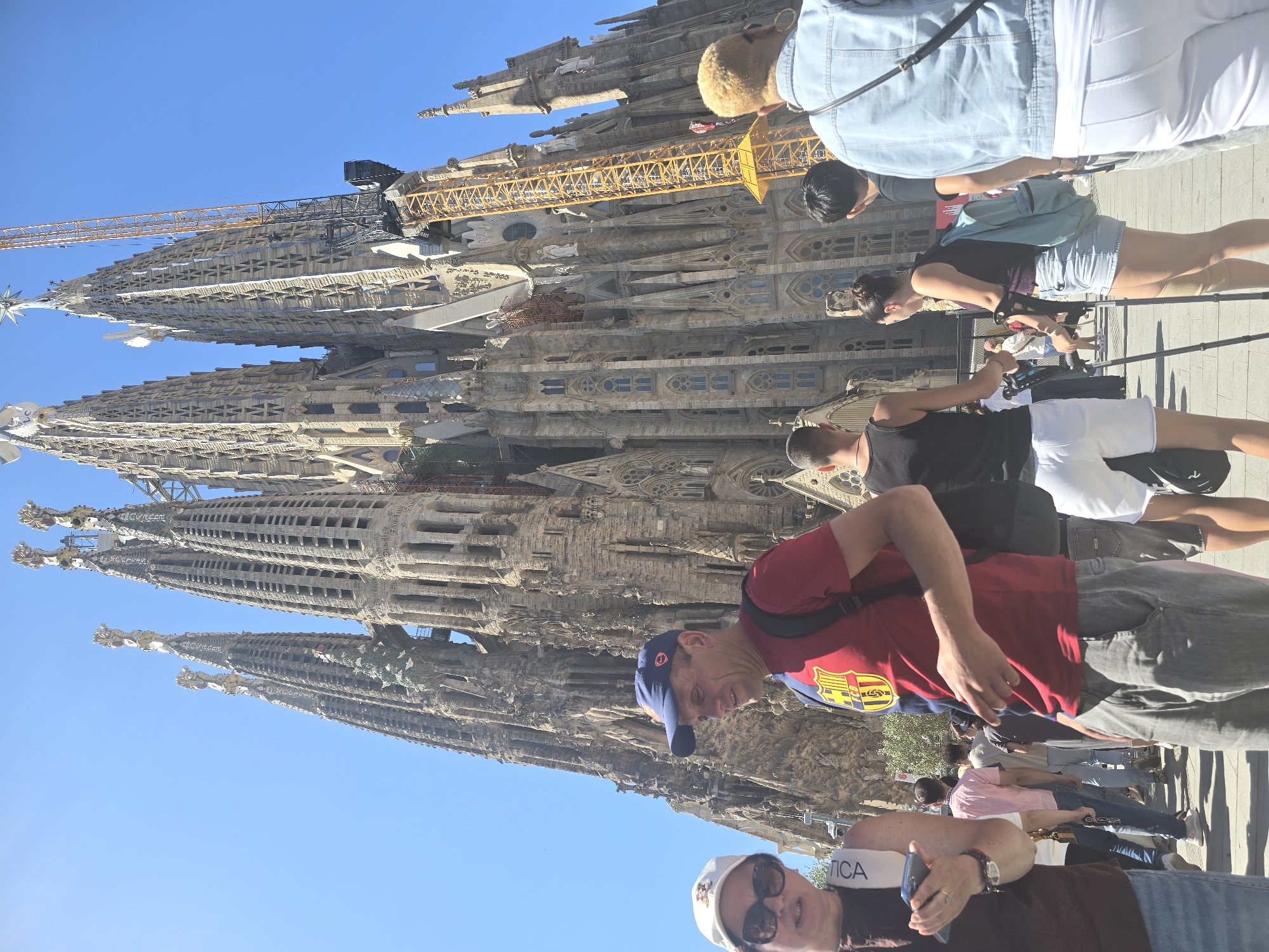 The image captures a sunny daytime view of the Sagrada Familia basilica in Barcelona, Spain. The iconic structure dominates the background with its intricate, spired architecture. A yellow construction crane is visible alongside one of the spires, indicating ongoing construction or renovation.
In the foreground, a diverse group of tourists is present. To the left, a woman wearing sunglasses and a white cap is looking towards the left side of the frame, holding a mobile phone. Next to her, a man in a red t-shirt with the FC Barcelona crest and a blue baseball cap is looking to his right. He is wearing grey pants and has a backpack on.
Further into the scene, other individuals are walking or standing. A person in a black tank top and white shorts is visible towards the center. To their right, a woman with a backpack, wearing a dark tank top and denim shorts, is using crutches. Other people, some with backpacks, are seen in the mid-ground. The ground appears to be paved with interlocking stones, common in pedestrian areas. The sky is clear and blue.