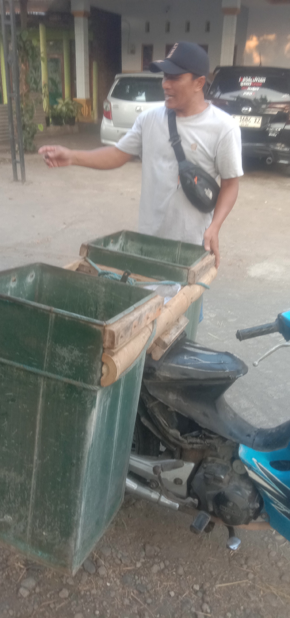 In Banjar Dowo, Indonesia, a man wearing a light-colored t-shirt, a black baseball cap, and a black fanny pack across his chest is standing next to a motorcycle. The motorcycle has a blue body and a silver exhaust pipe. Attached to the back of the motorcycle is a wooden frame holding two green, rectangular bins. The man is gesturing with his left hand. In the background, there are two cars and a building with a covered porch. The ground is a mix of dirt and gravel.