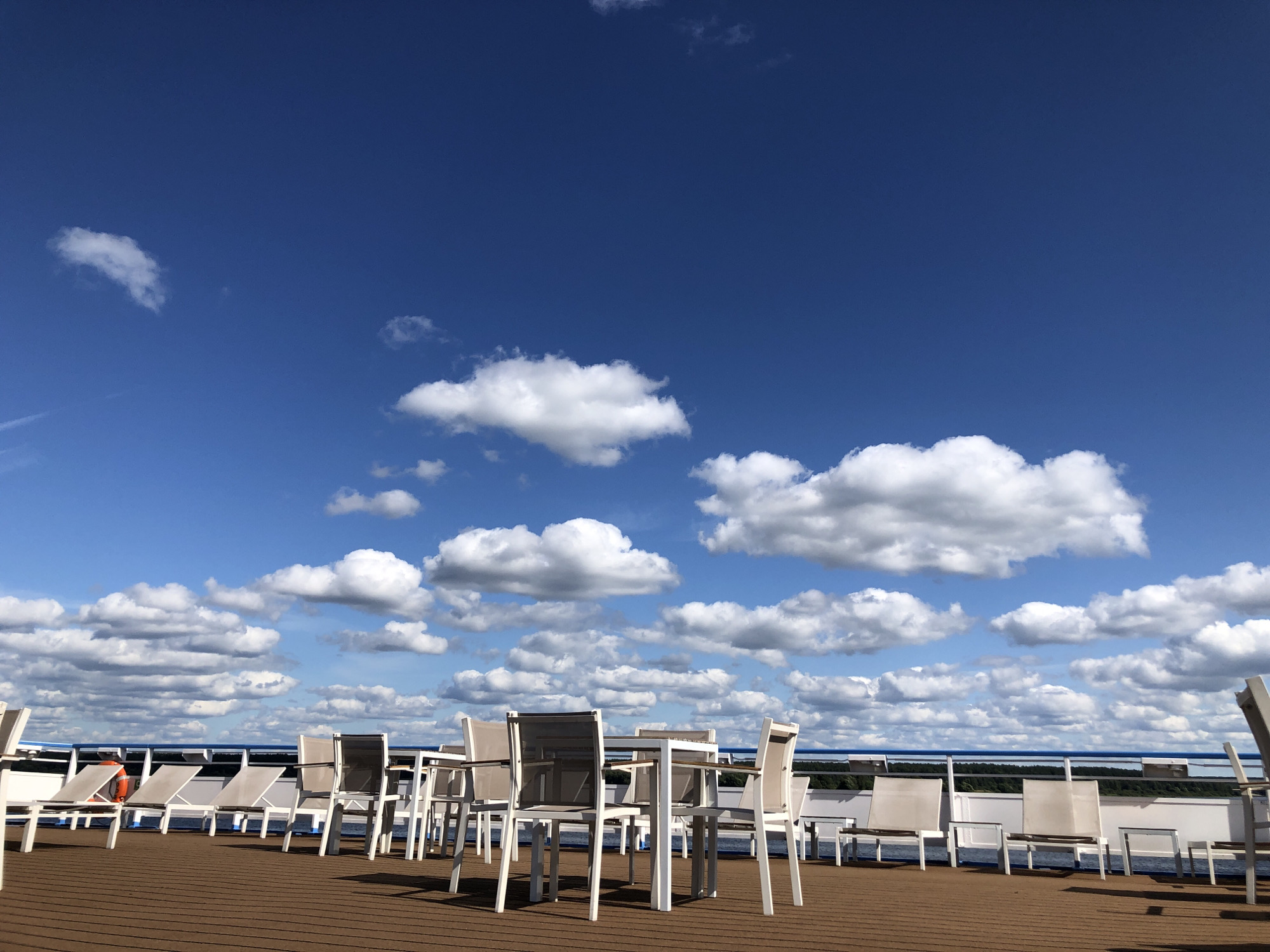 The image depicts an outdoor seating area on a wooden deck, likely a cruise ship or waterfront establishment. Numerous empty chairs and tables are arranged, suggesting a place for relaxation or dining. The background features a bright blue sky dotted with fluffy white clouds. Beyond the deck, a railing overlooks a body of water, with a distant treeline visible. A red life preserver is attached to the railing. The scene is peaceful and devoid of people. The location is stated to be in the city of Myshkin, Russia.